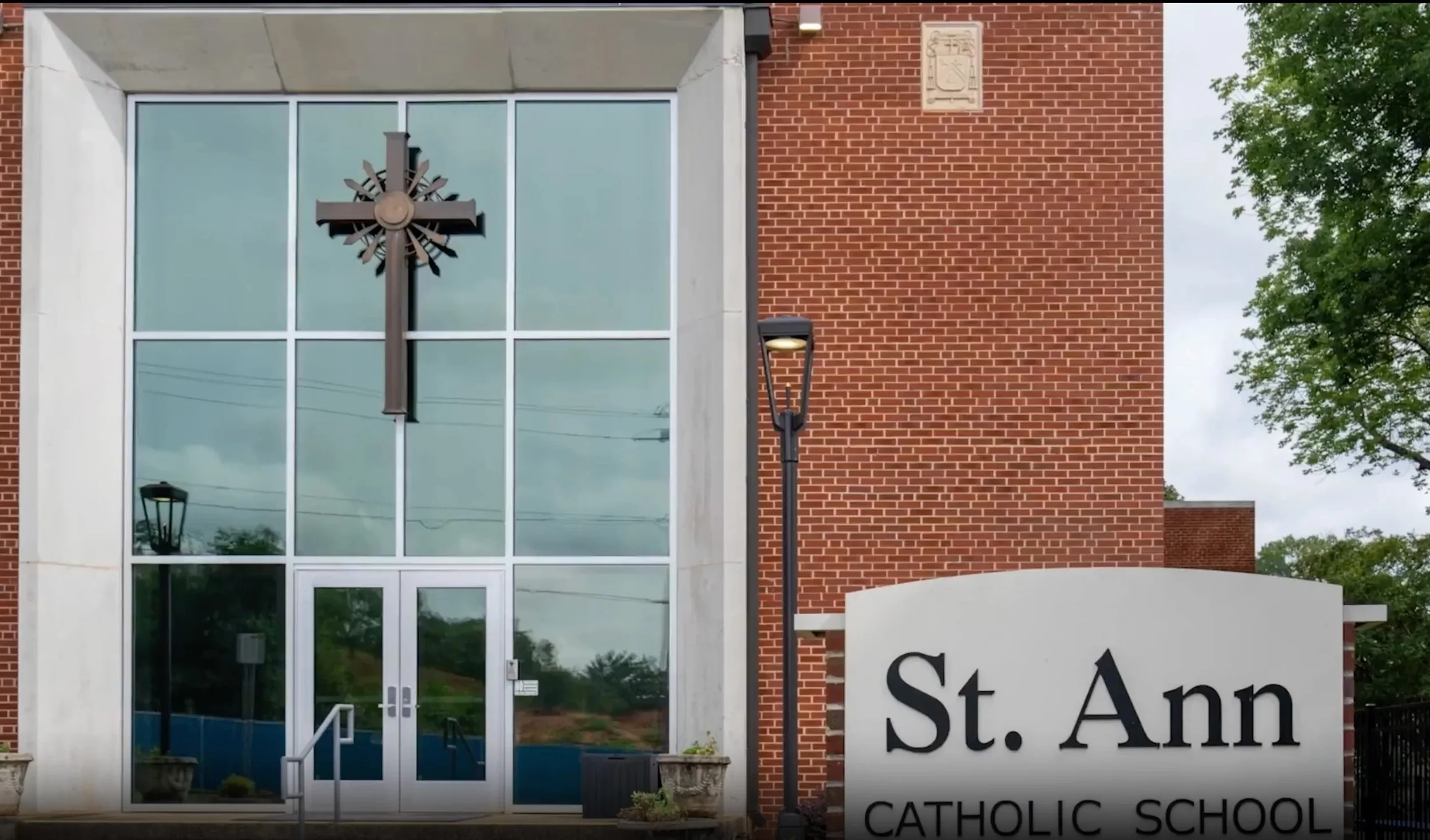 Exterior of a building with a large glass window and a brick wall, featuring a cross monument in front, and a sign that reads 'St. Ann Catholic School'.