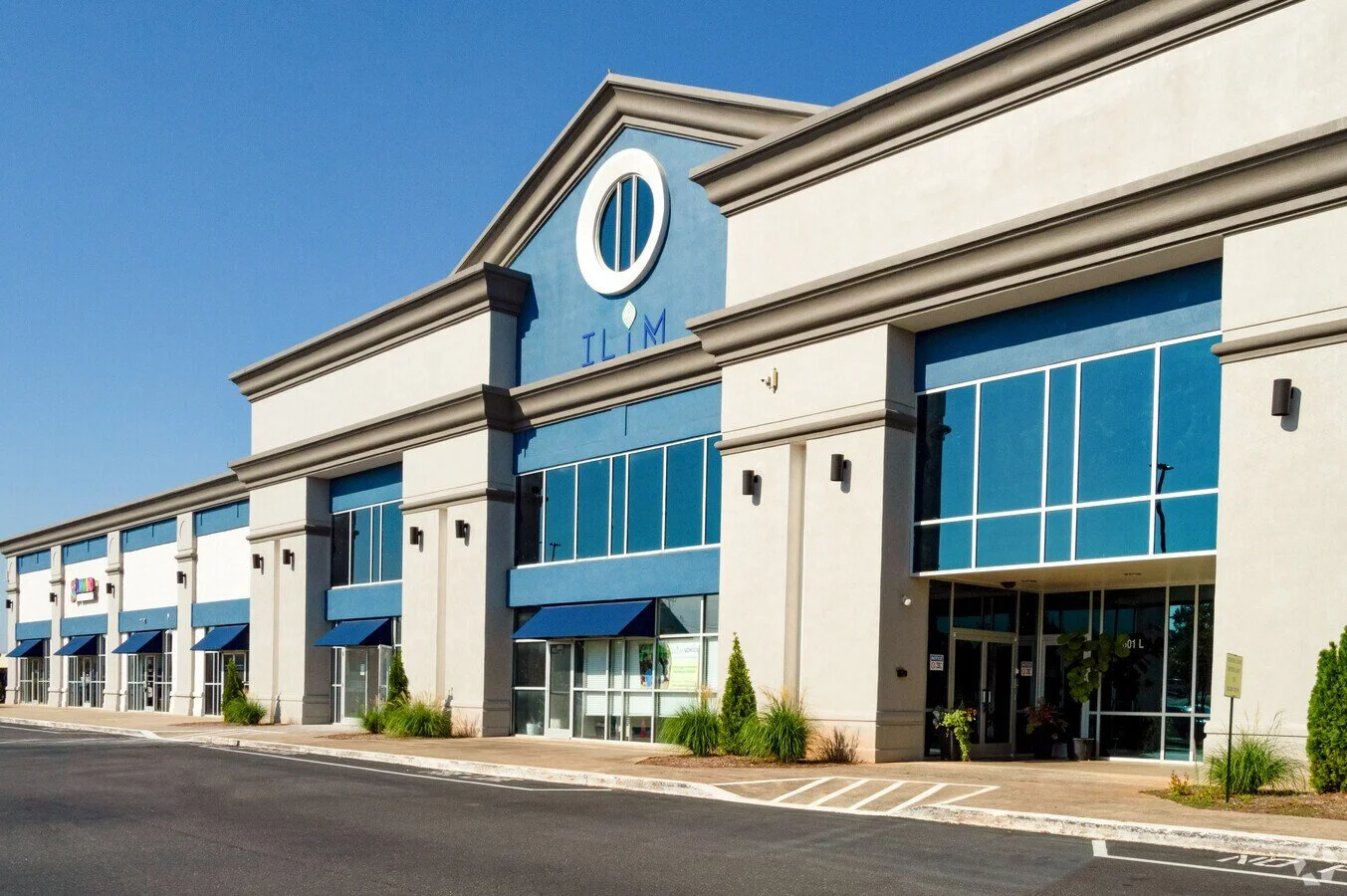 Exterior view of a modern shopping center with retail stores and large glass windows, blue accents, and well-maintained landscaping.