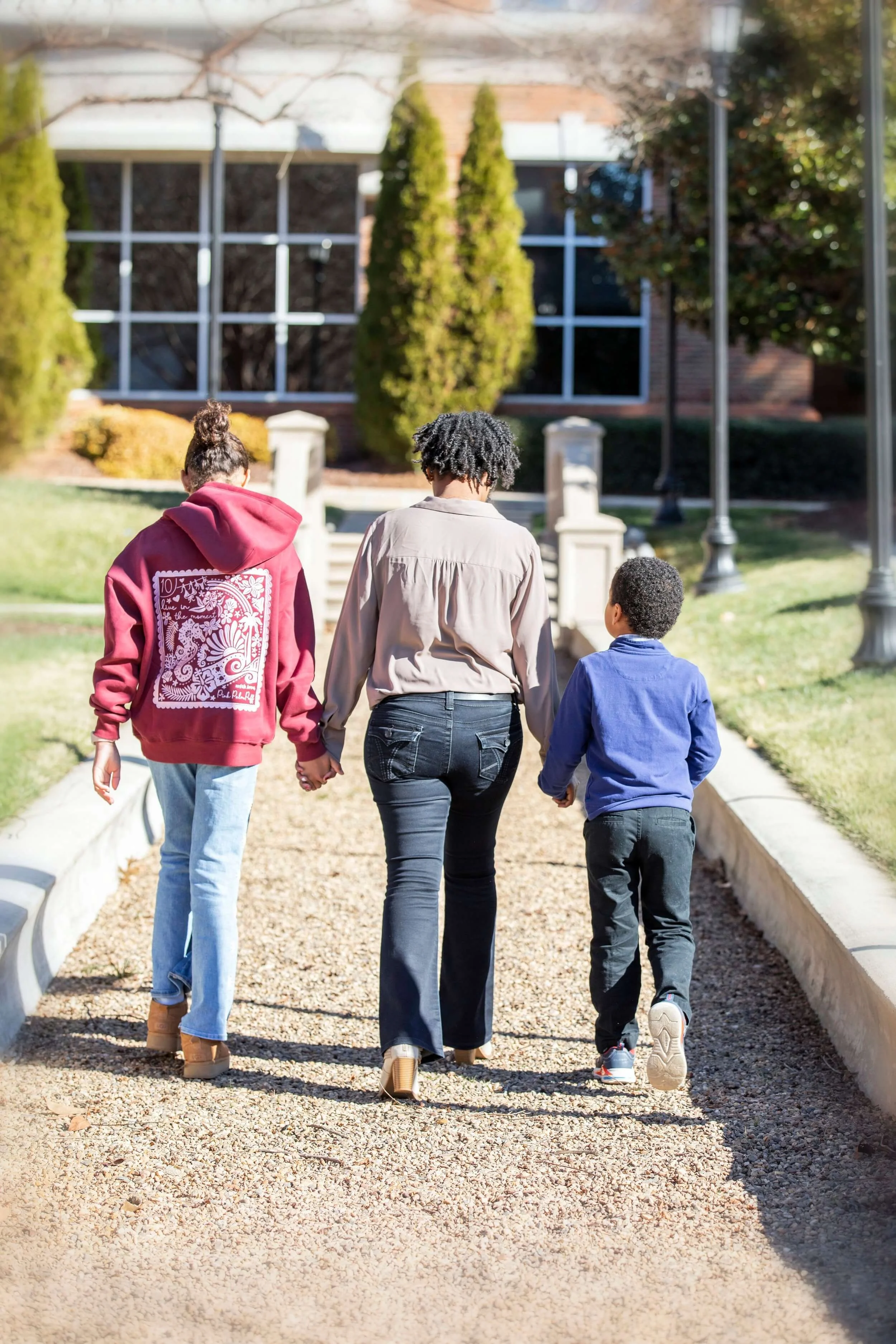 Three people, an adult woman and two children, walking hand-in-hand on a gravel path outside near a brick building, surrounded by trees and greenery.