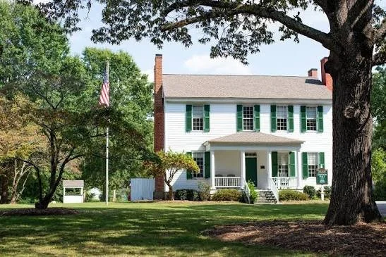 A large white two-story house with green shutters and a front porch, surrounded by trees and a well-maintained lawn, American flag flying on a flagpole nearby.