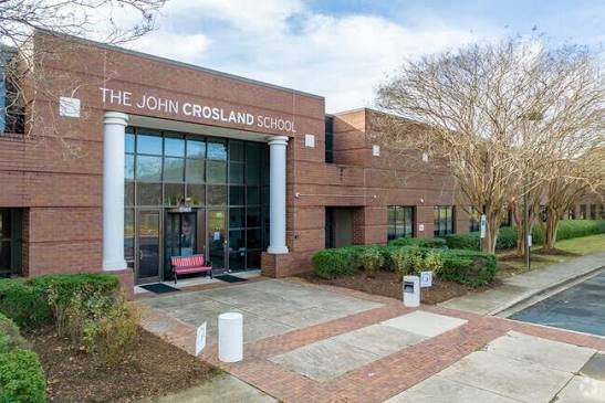 Exterior view of The John Crosland School building with glass entrance, red brick walls, white columns, benches, shrubs, and leafless trees.