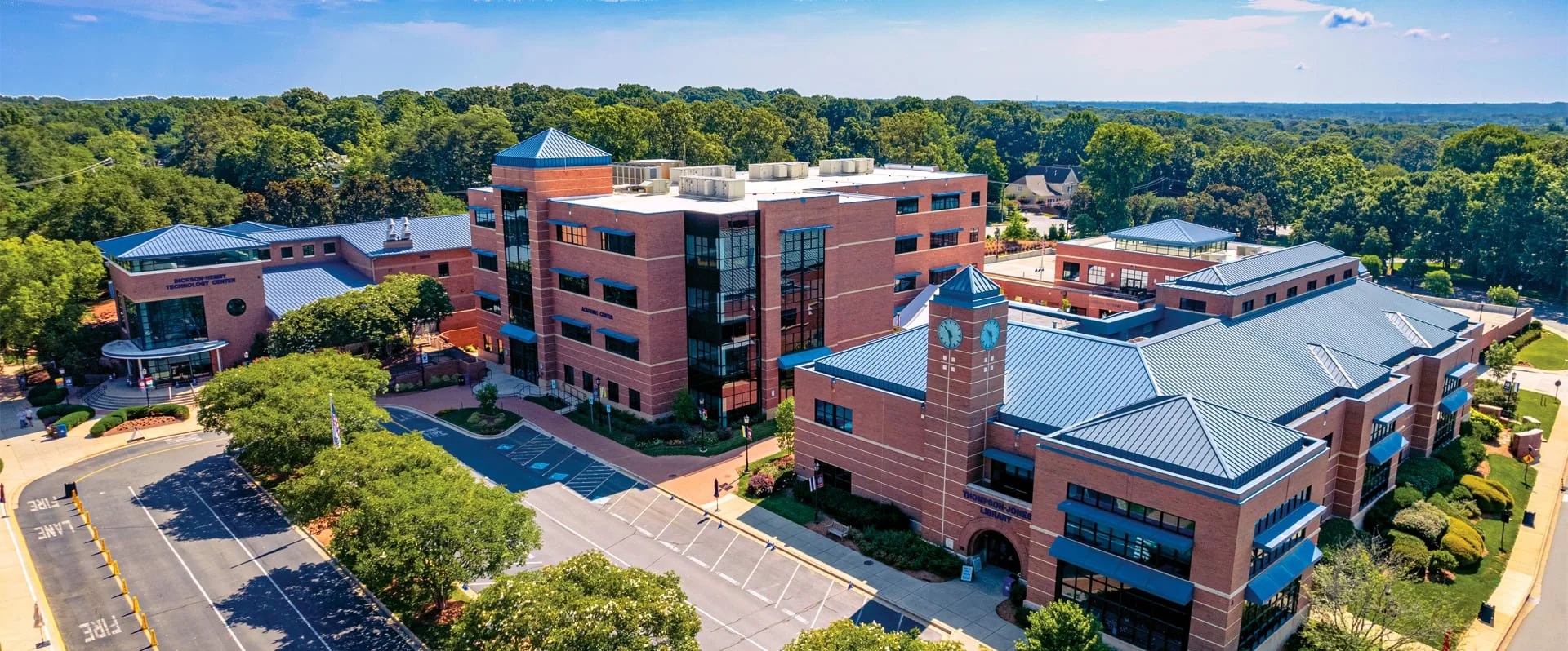 Aerial view of a large brick academic building with a blue metal roof, clock tower, and surrounding parking lot, greenery, and trees.