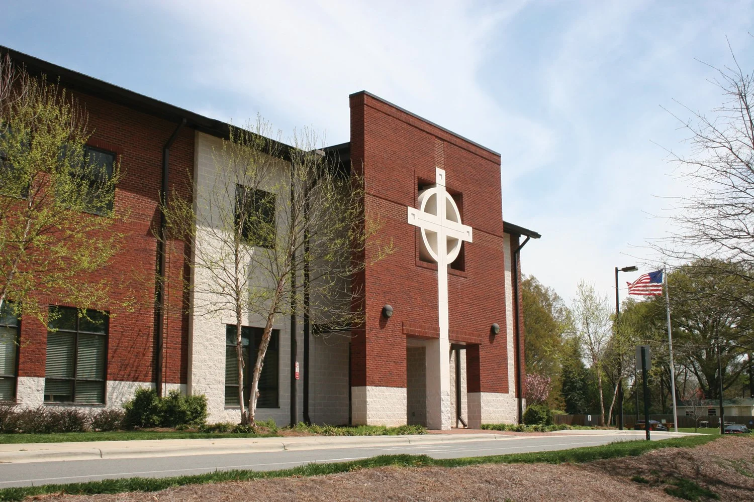Modern church building with a large white cross mounted on a red brick wall, trees with budding leaves, and an American flag on a flagpole outside.