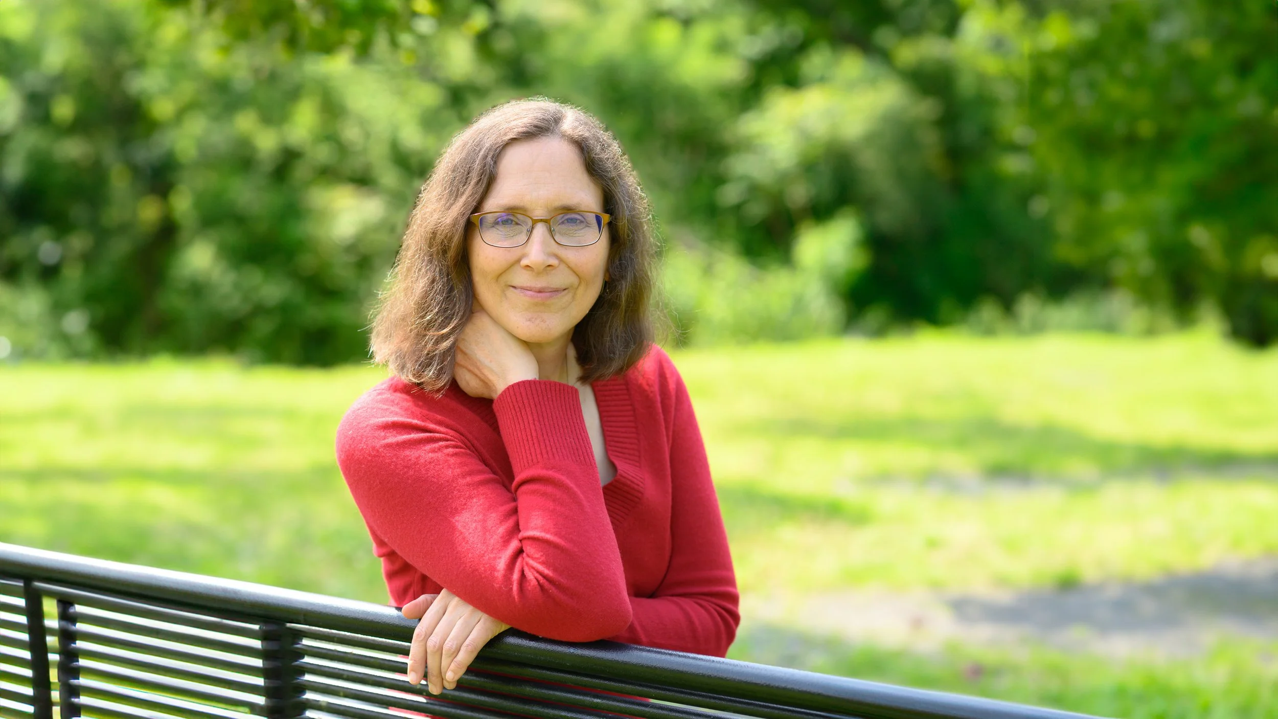 A woman with glasses and shoulder-length hair wearing a red sweater sitting on a park bench in a green outdoor setting.