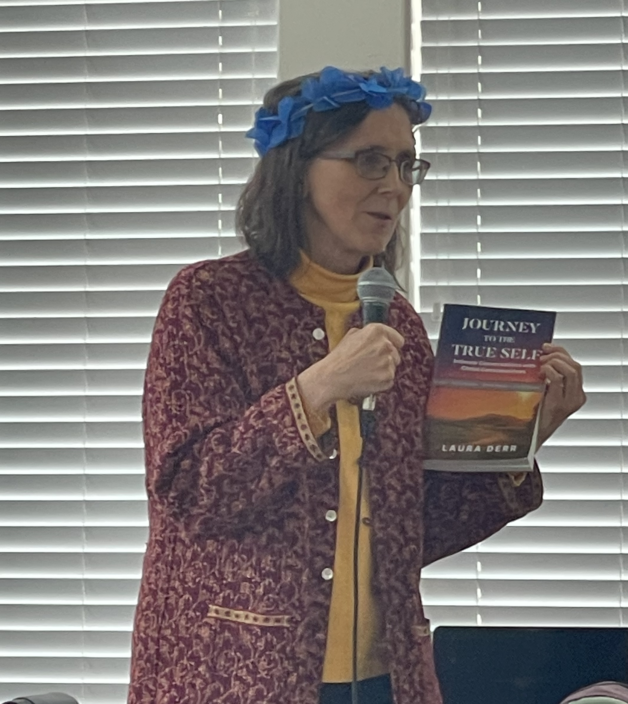 A woman with glasses, wearing a blue ribbon headband, speaking into a microphone, holding a book titled 'Journey to the True Self'. She stands in front of closed horizontal blinds.