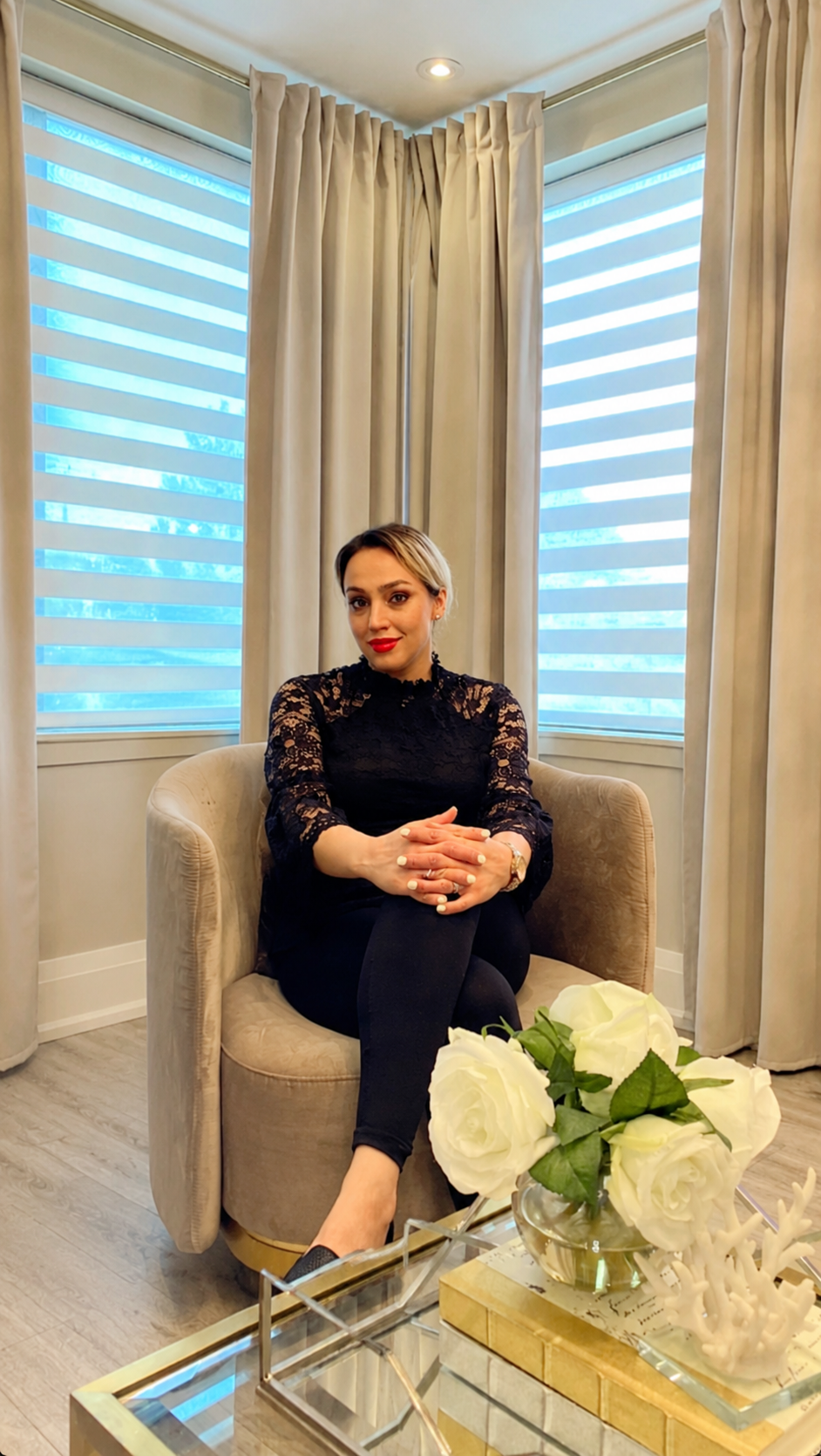 A woman sitting on a beige armchair with her legs crossed, wearing a black lace top and black pants, in a well-lit room with large windows and beige curtains, with a glass table in front holding a vase of white roses.