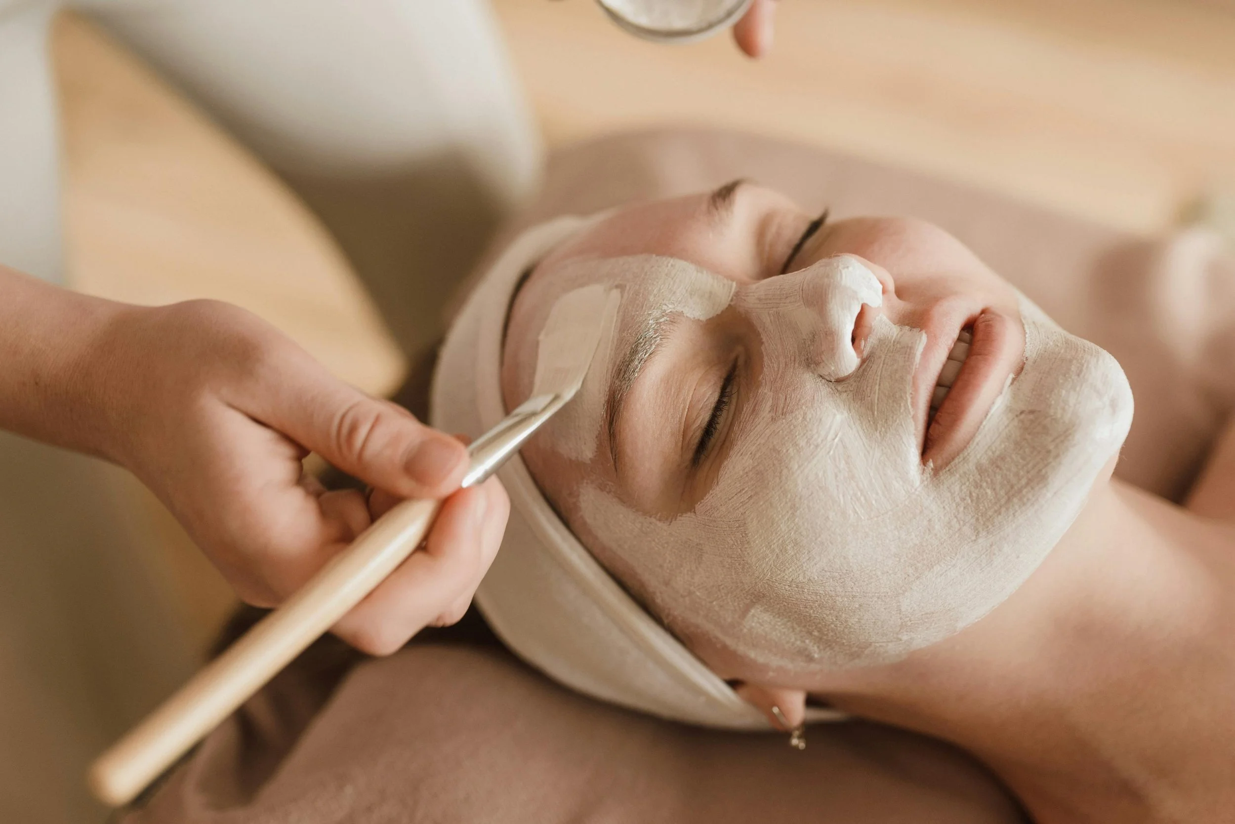 A person receiving a facial facial treatment with a brush, while lying down with eyes closed. The face is covered with a white facial mask, and a skincare professional applies the mask with a brush.