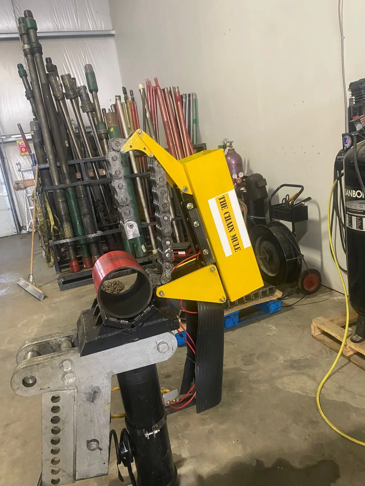A large robot with a yellow frame and black components stands inside a garage or workshop. It has a rolling structure with many metal rods and parts stored in racks behind it, and a chain is visible on the side. The scene includes various tools and e