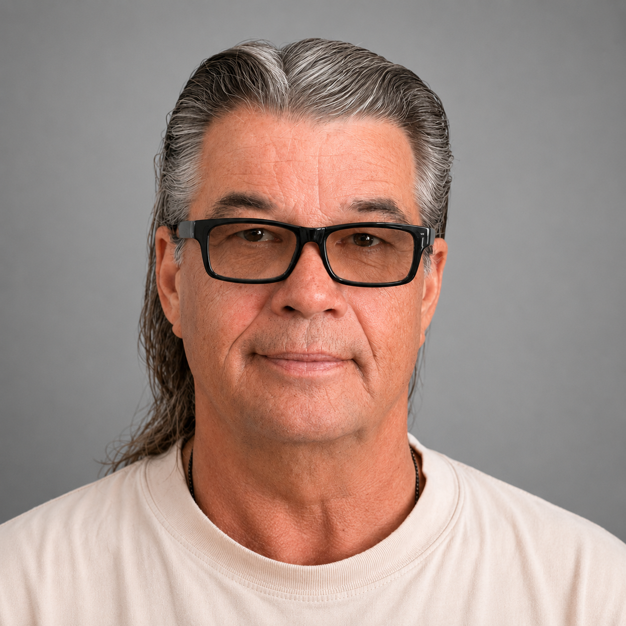 Portrait of a middle-aged man with long gray hair, wearing black glasses and a light-colored shirt, smiling gently against a plain gray background.