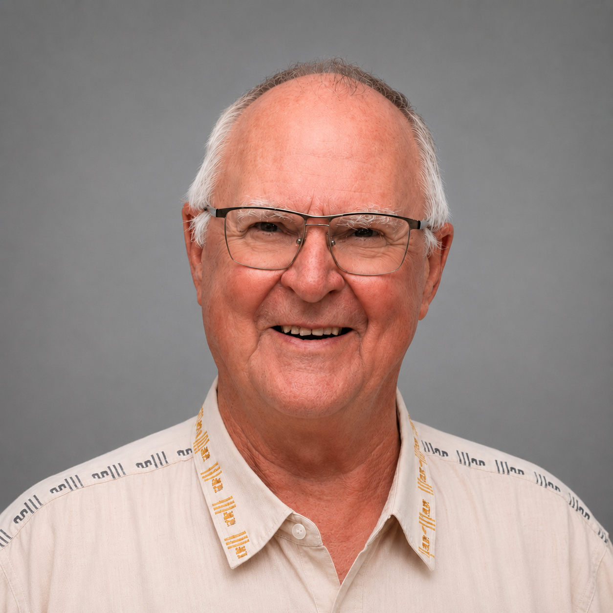 Portrait of an elderly man with glasses smiling, wearing a beige shirt with a collar and lanyard, against a gray background.