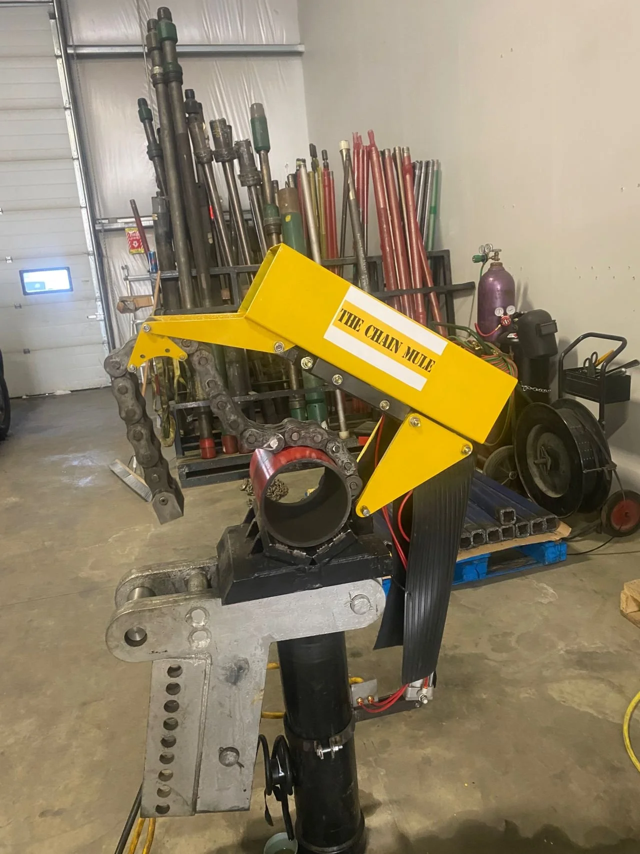 A hydraulic chain saw machine labeled "The Chain Mule" set up in a warehouse with metal pipes, a gas cylinder, and equipment in the background.