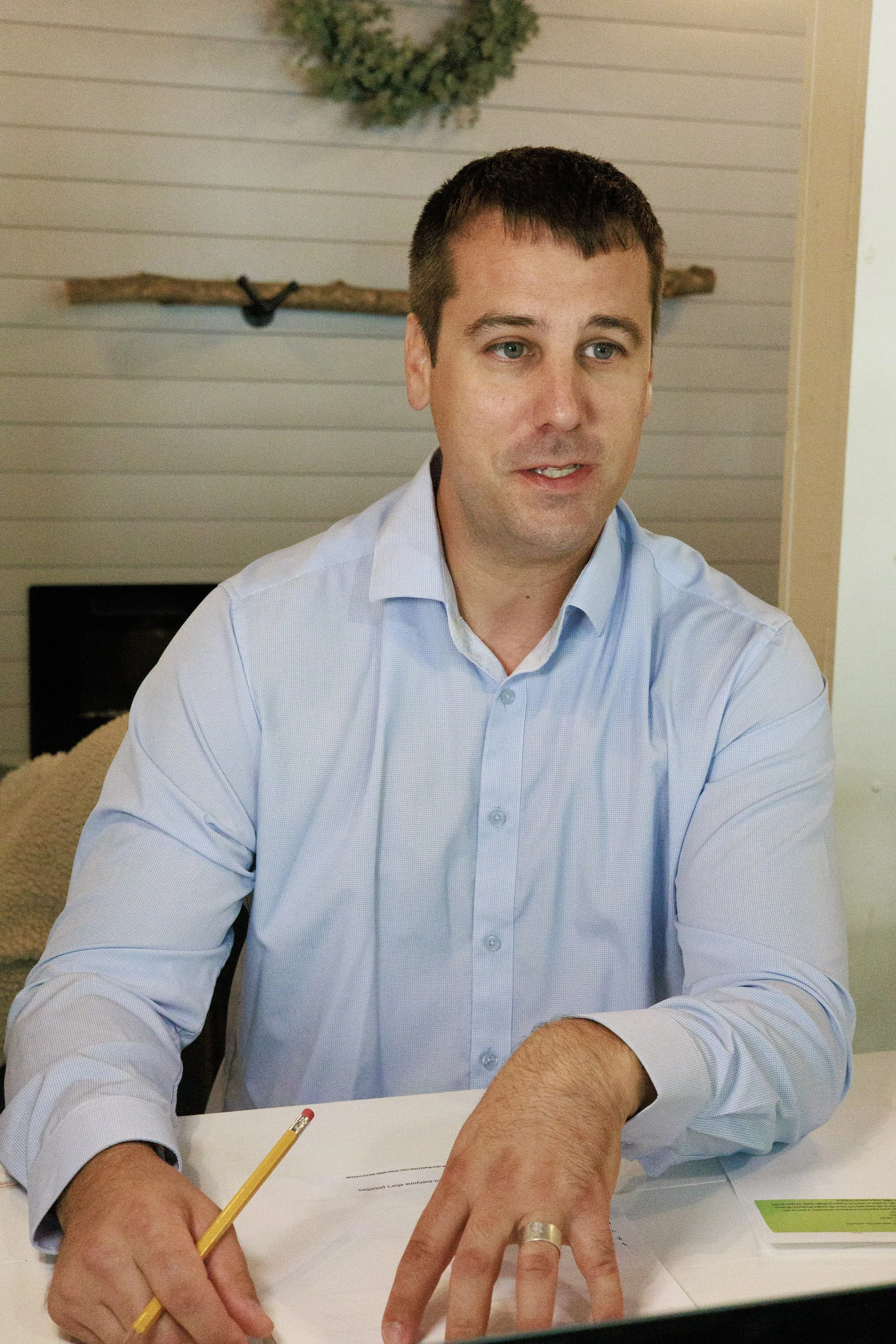 A man with short dark hair and light skin wearing a light blue button-up shirt, is sitting at a desk. He is holding a yellow pencil and showing both hands, with a ring on his left ring finger. The background includes a decorative wall with a wooden branch and a green wreath above.