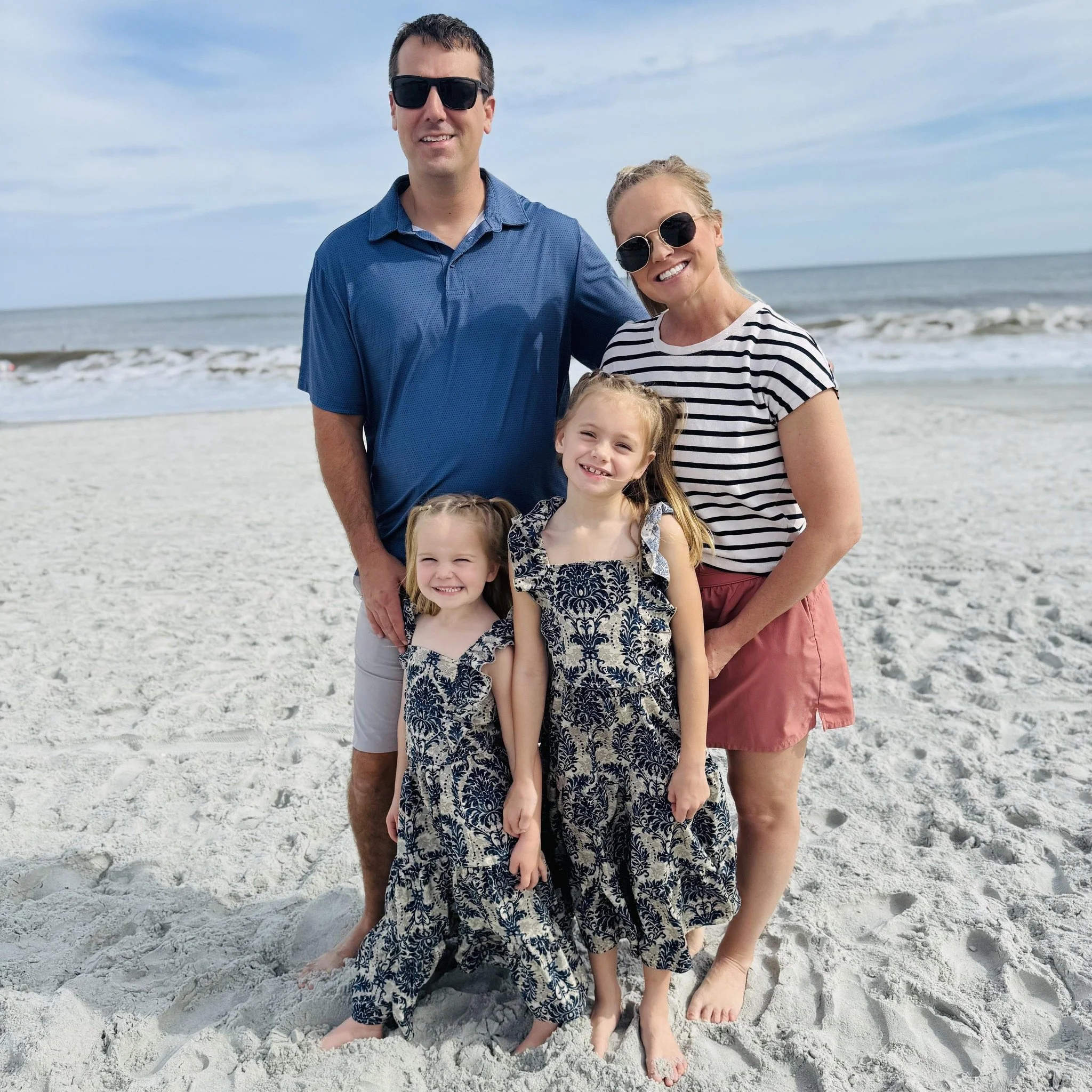 Family of five at the beach, with waves in the background, smiling, wearing sunglasses, and standing on sand.