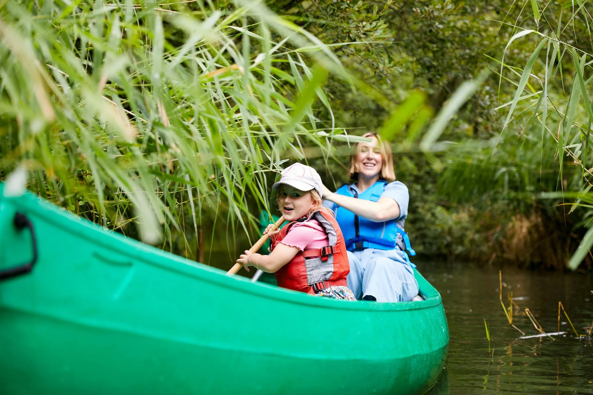 Two people, a woman and a young girl, are paddling a green kayak through a narrow waterway surrounded by tall green grass and bushes, enjoying an outdoor adventure.