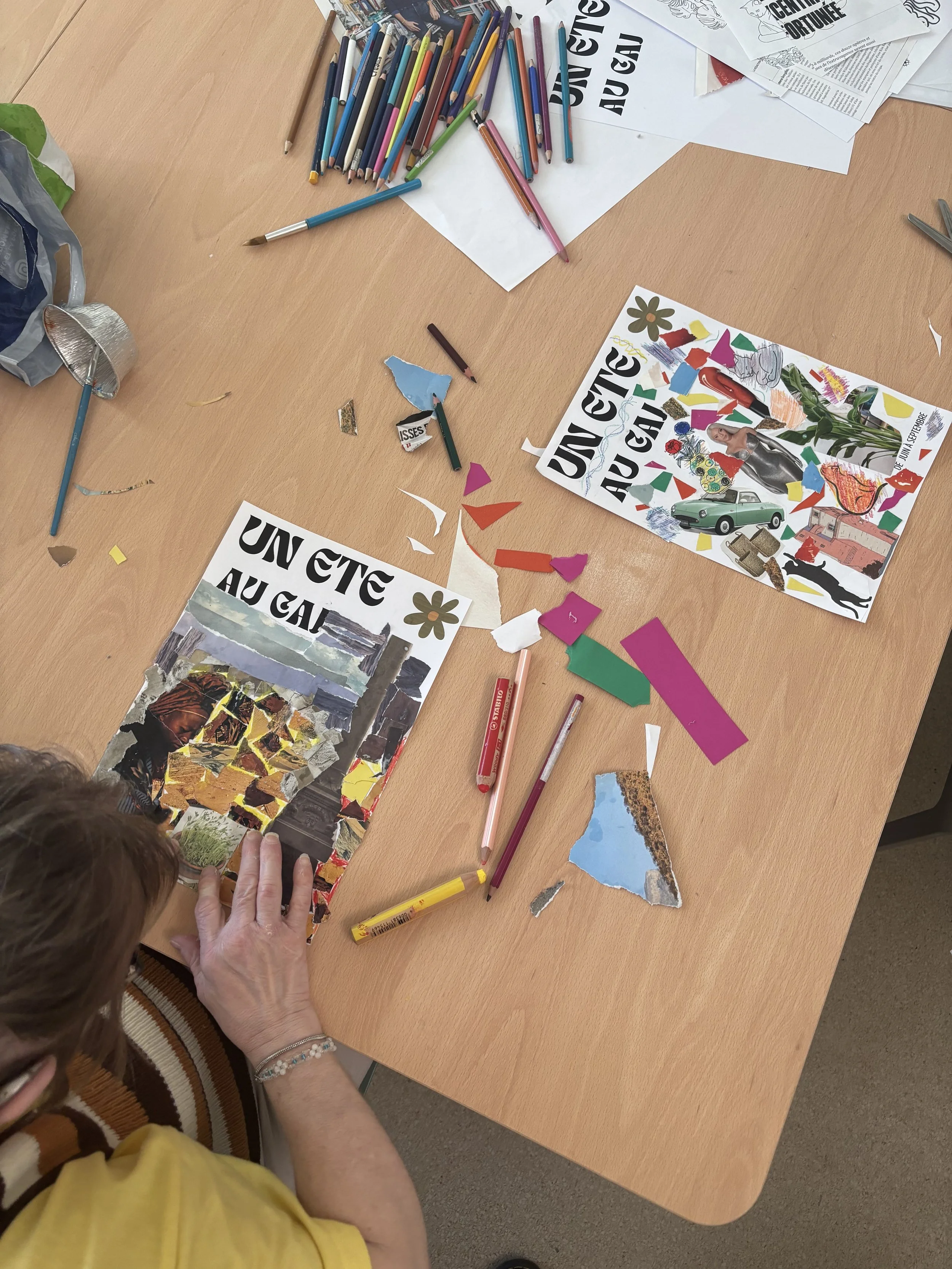 A person with short hair and wearing a yellow shirt is collage-making, placing a torn piece of collage artwork on a table. The table has colored pencils, torn paper, and collage materials scattered on it.