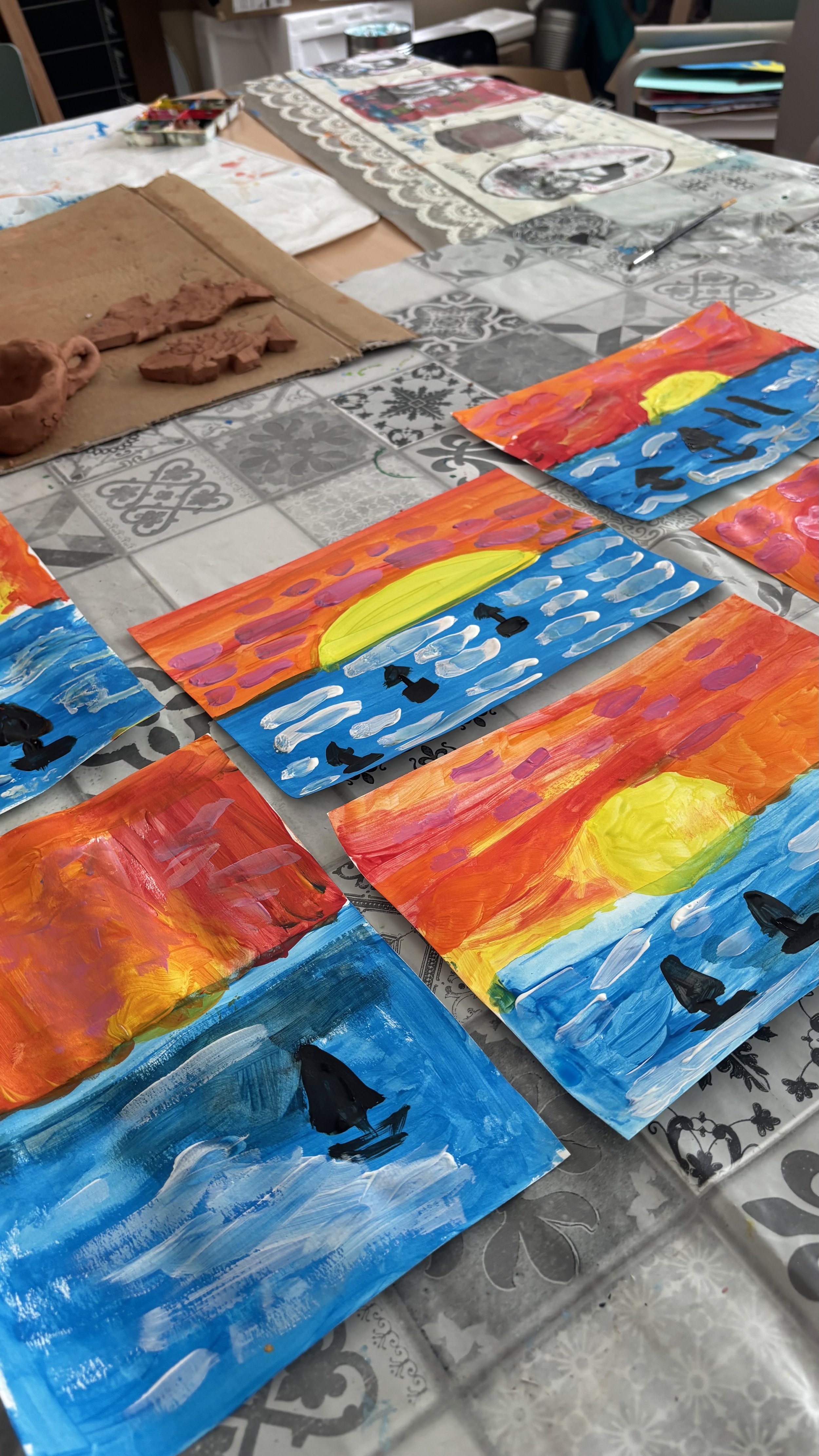 Multiple small paintings of sunsets over water with silhouetted boats, painted with bright colors and expressive brushstrokes, displayed on a table in an art classroom.