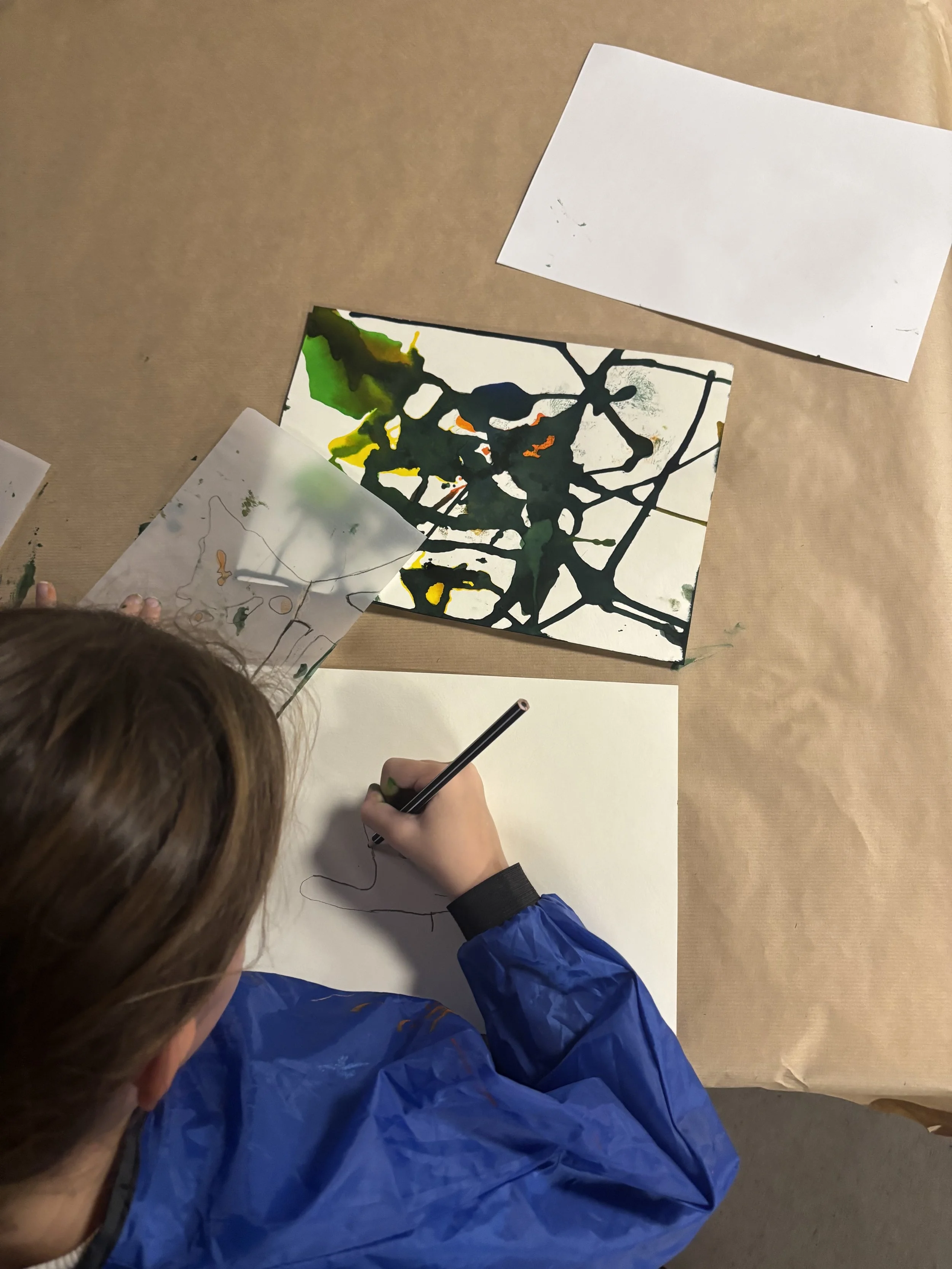 A person with brown hair wearing a blue jacket is sitting at a table, drawing on a piece of paper with a pen or pencil. There are other papers with colorful abstract or splatter designs on the table.