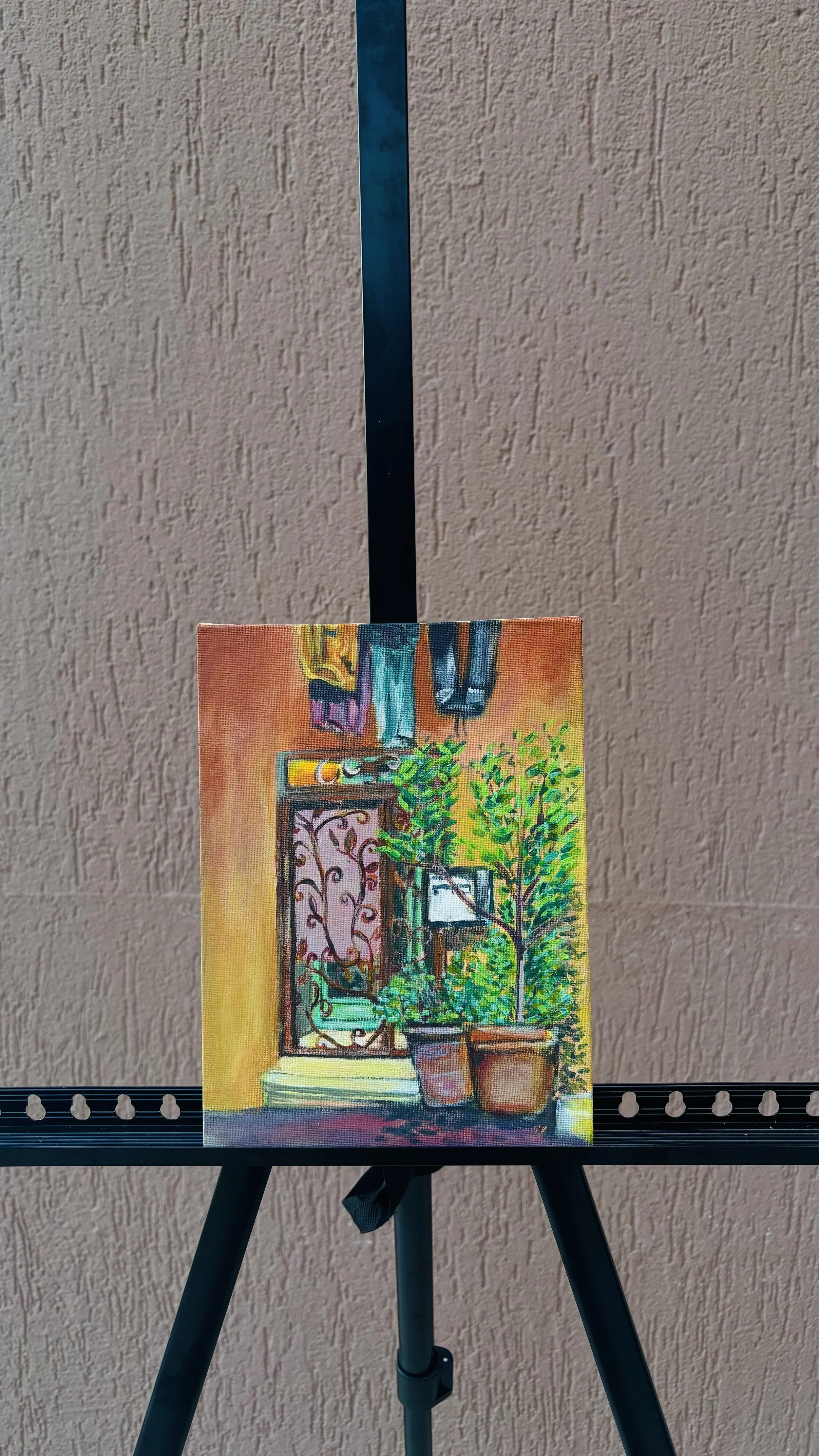 A small colorful painting on an easel depicts a window with a view of green trees and plants outside, with laundry hanging above and a beige wall in the background.