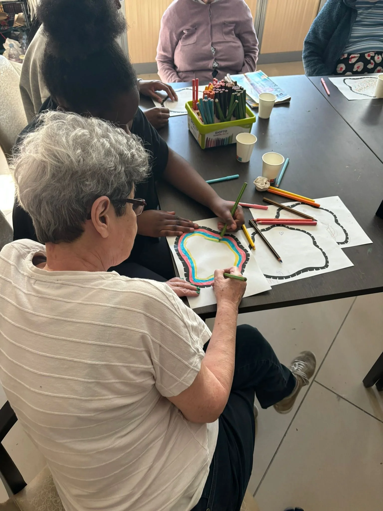 People seated around a table coloring pictures of rainbows and clouds with colored pencils in an indoor setting.