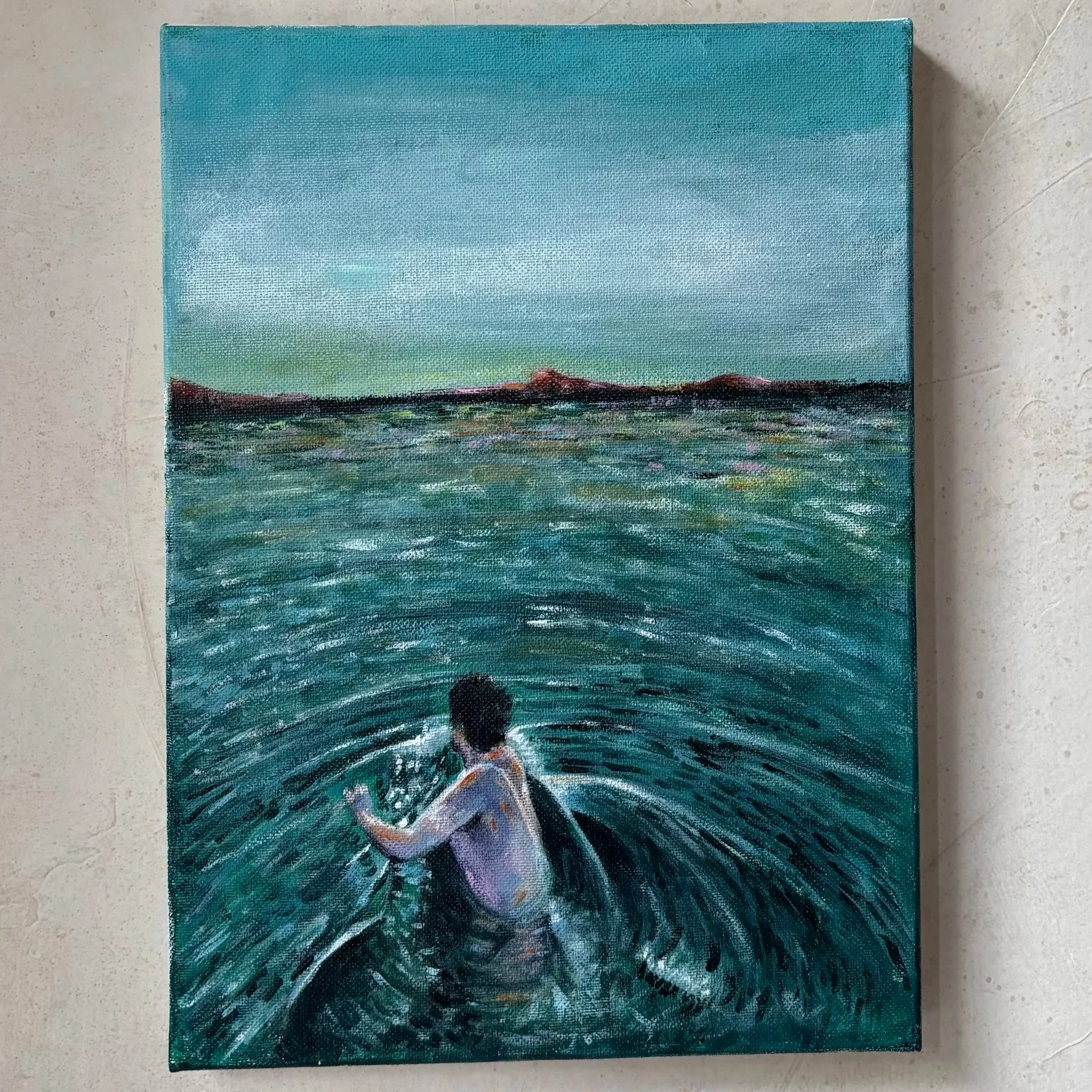 A small acrylic painting of a landscape scene with a blue sky, distant mountains, and a body of water in the foreground, resting on a beige tiled floor.
