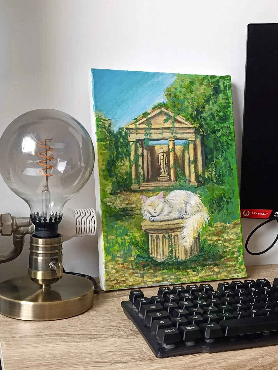 A painting of a classical building with columns and a statue in the background, surrounded by greenery, depicts a resting white cat on a pedestal in front of the building, on a wooden surface next to a modern light bulb lamp and a computer monitor.
