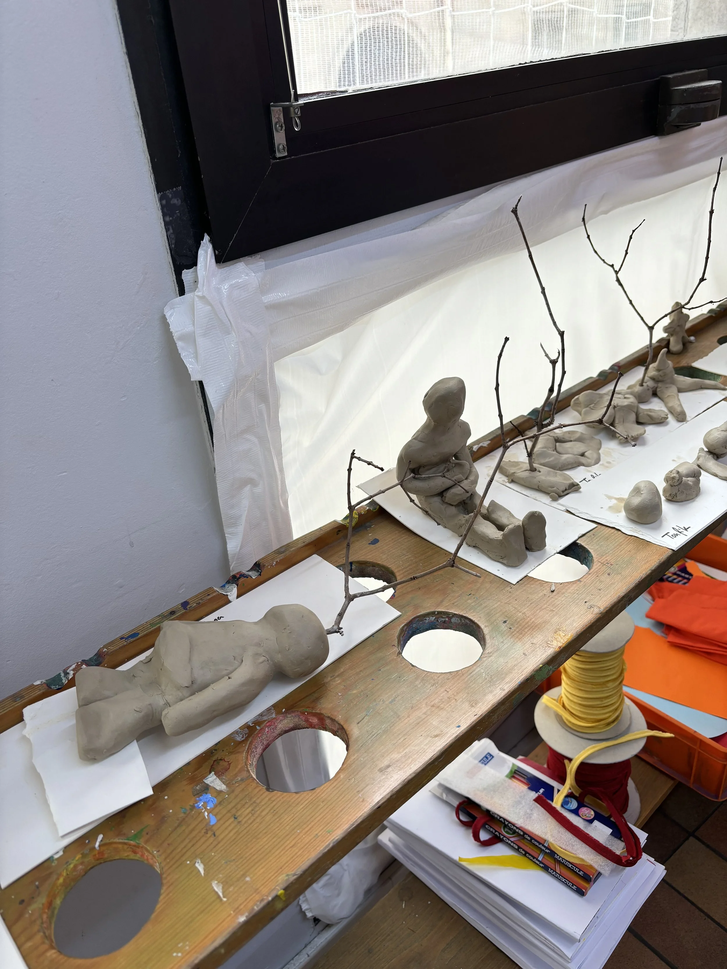 Clay sculptures and sketches of people, trees, and objects on a workspace table with art supplies.