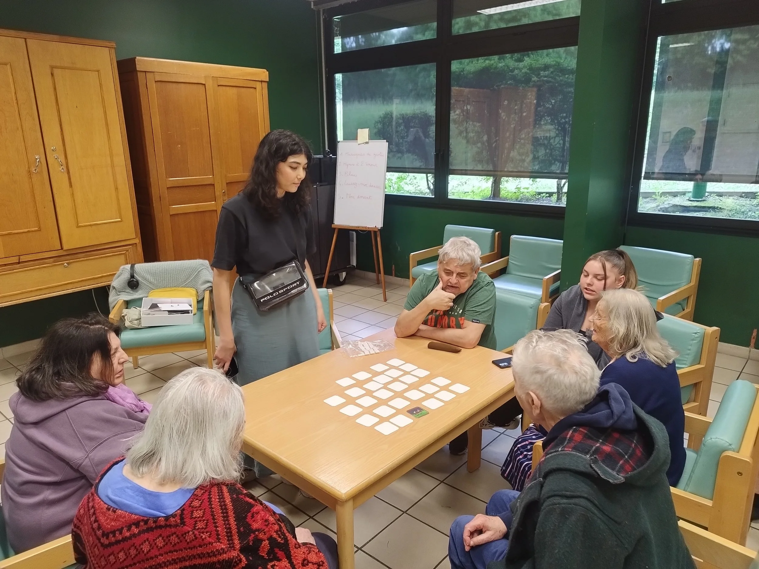 A woman standing in a room with several elderly people seated at a table, engaging in conversation or a group activity, with a whiteboard and large windows in the background.