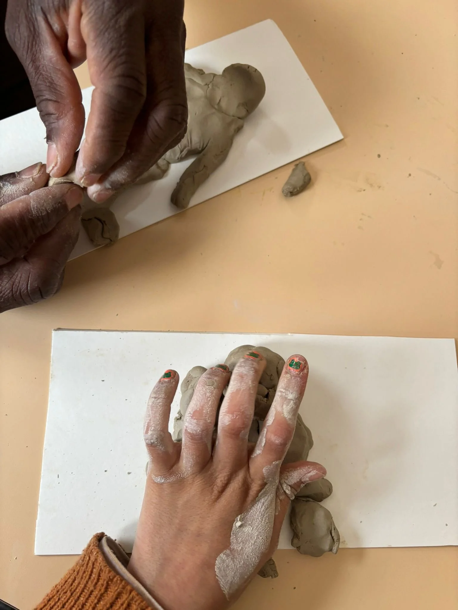 Two hands shaping gray modeling clay on white paper, with additional clay pieces nearby on a beige surface.