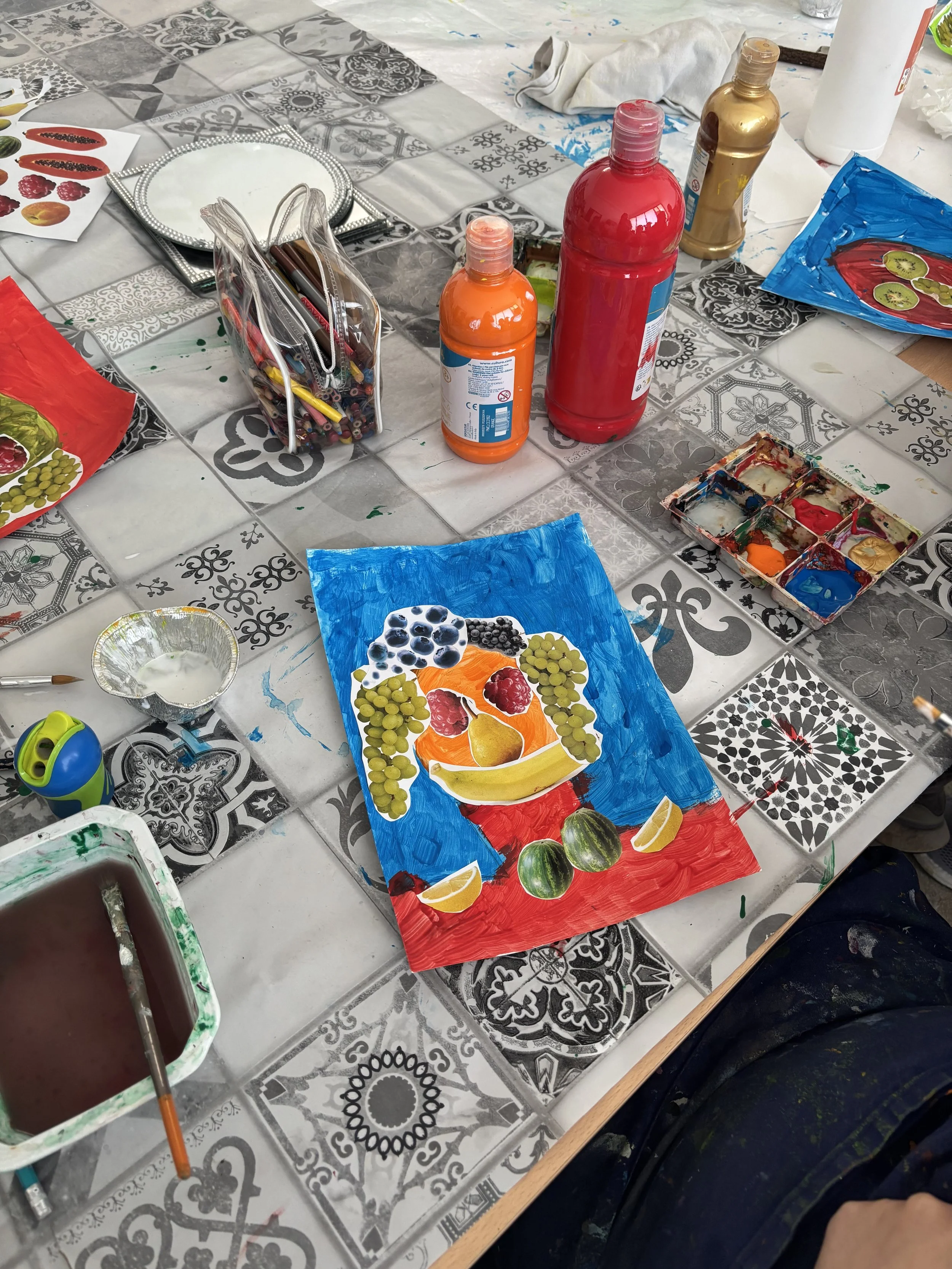 Table covered with art supplies including bottles of paint, a palette, a partly painted poster of fruit, and various tools for painting.