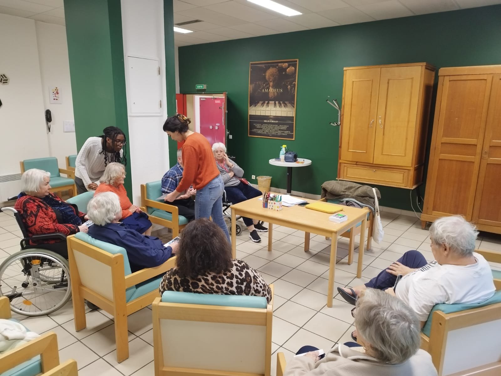 Older adults sitting in a room, some in wheelchairs, engaging with caregivers or volunteers during a group activity or social event.