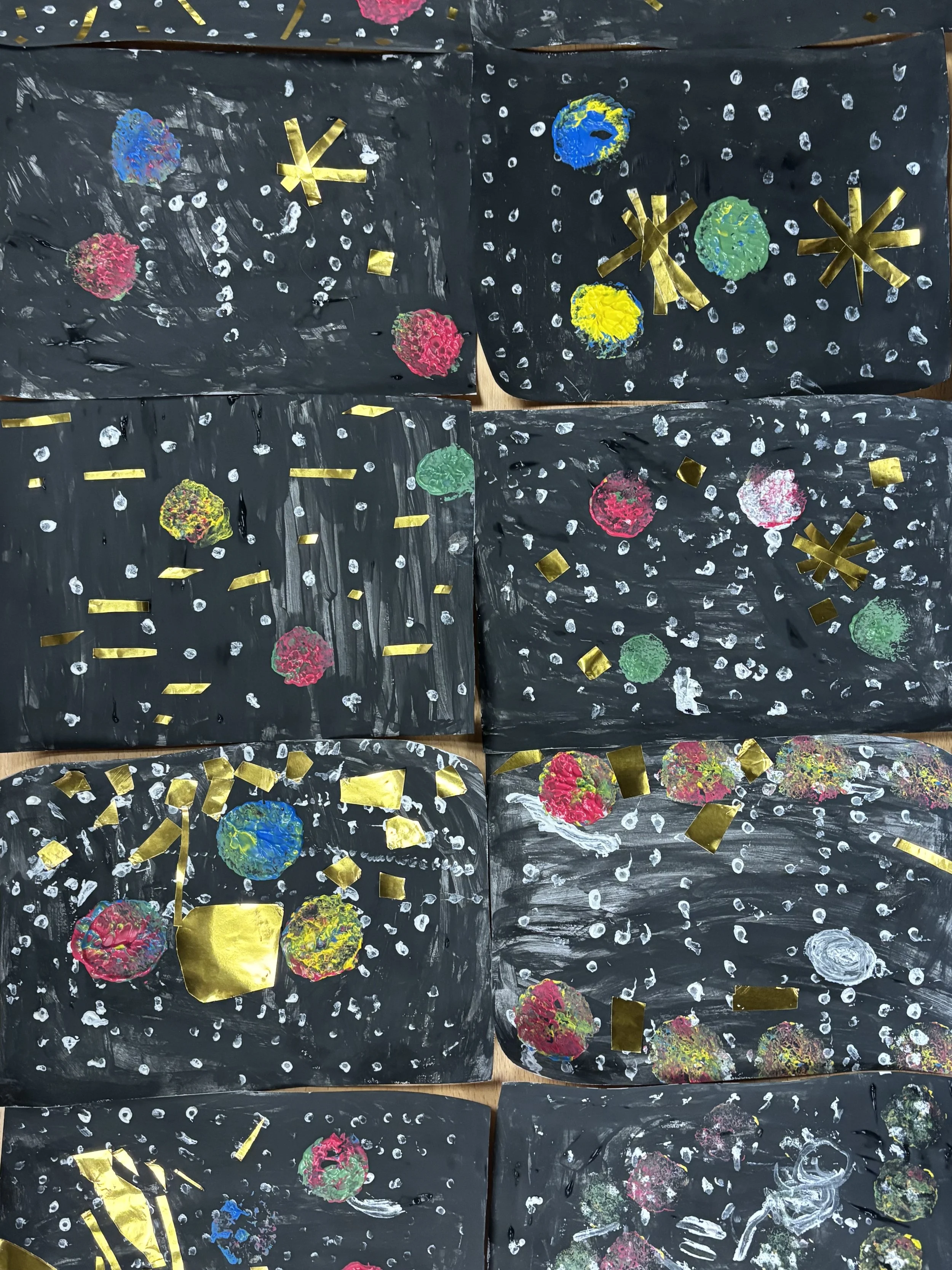 A creative space craft painting using black paper as background, decorated with colorful painted planets, white dots representing stars, and cut-out gold paper shapes resembling stars and geometric patterns.