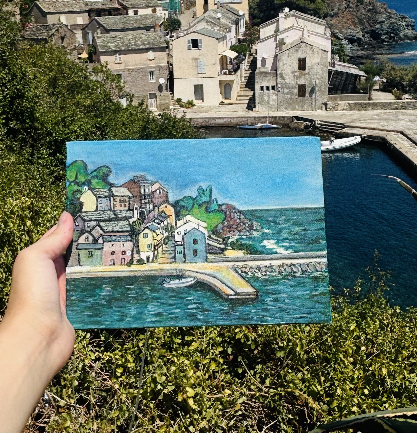Person holding a painting of a coastal village, with similar houses, boats, and water as in the background.