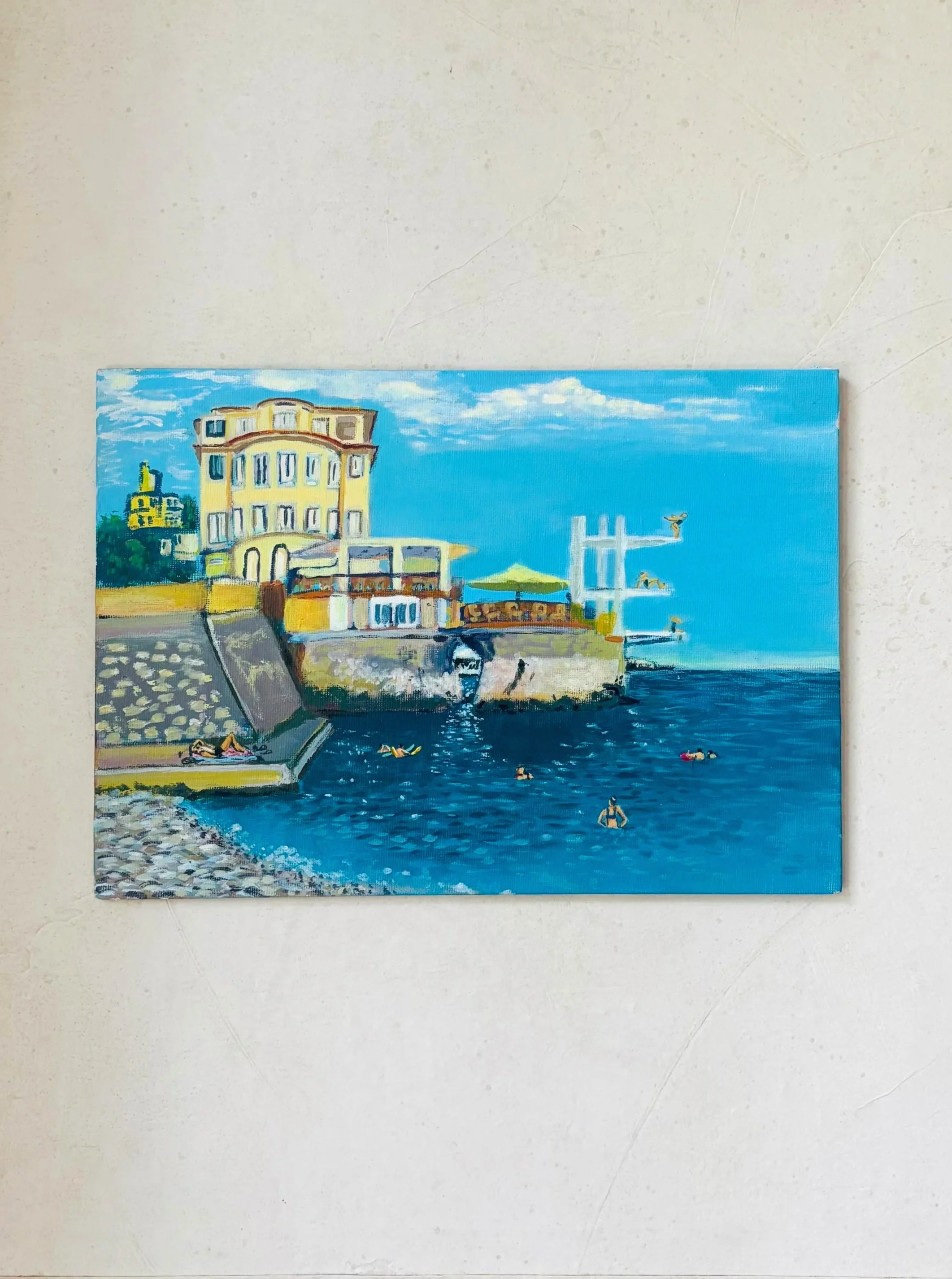 A painting of a seaside scene with a large yellow building, a pebble beach, people swimming, and a pier extending into the water, under a blue sky with clouds.