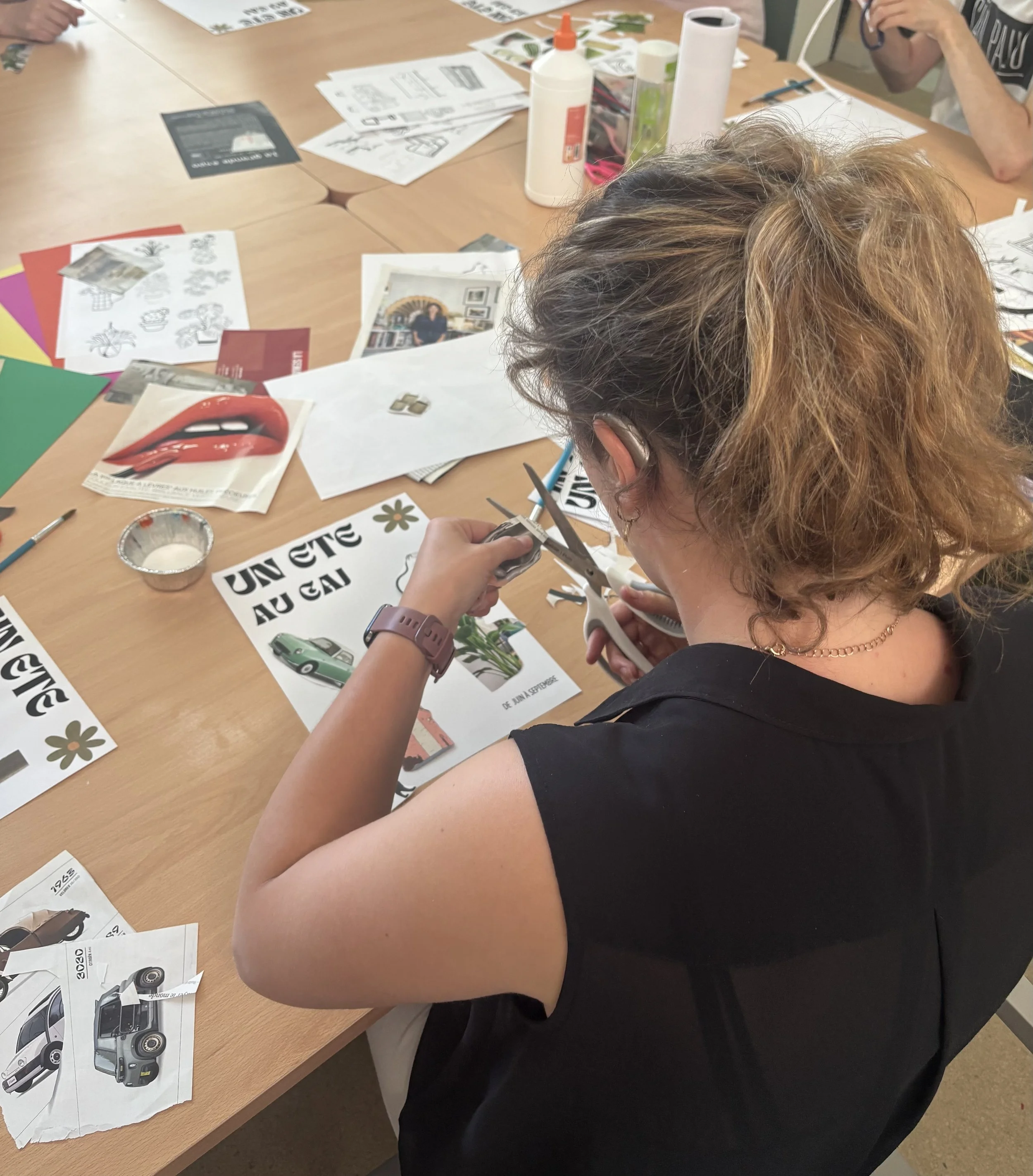 A person with light brown and blonde curly hair, wearing a black sleeveless top and gold chain necklace, is cutting paper with scissors on a table. The table has various printed materials, including car brochures, flyers, glue, tape, and tissue paper