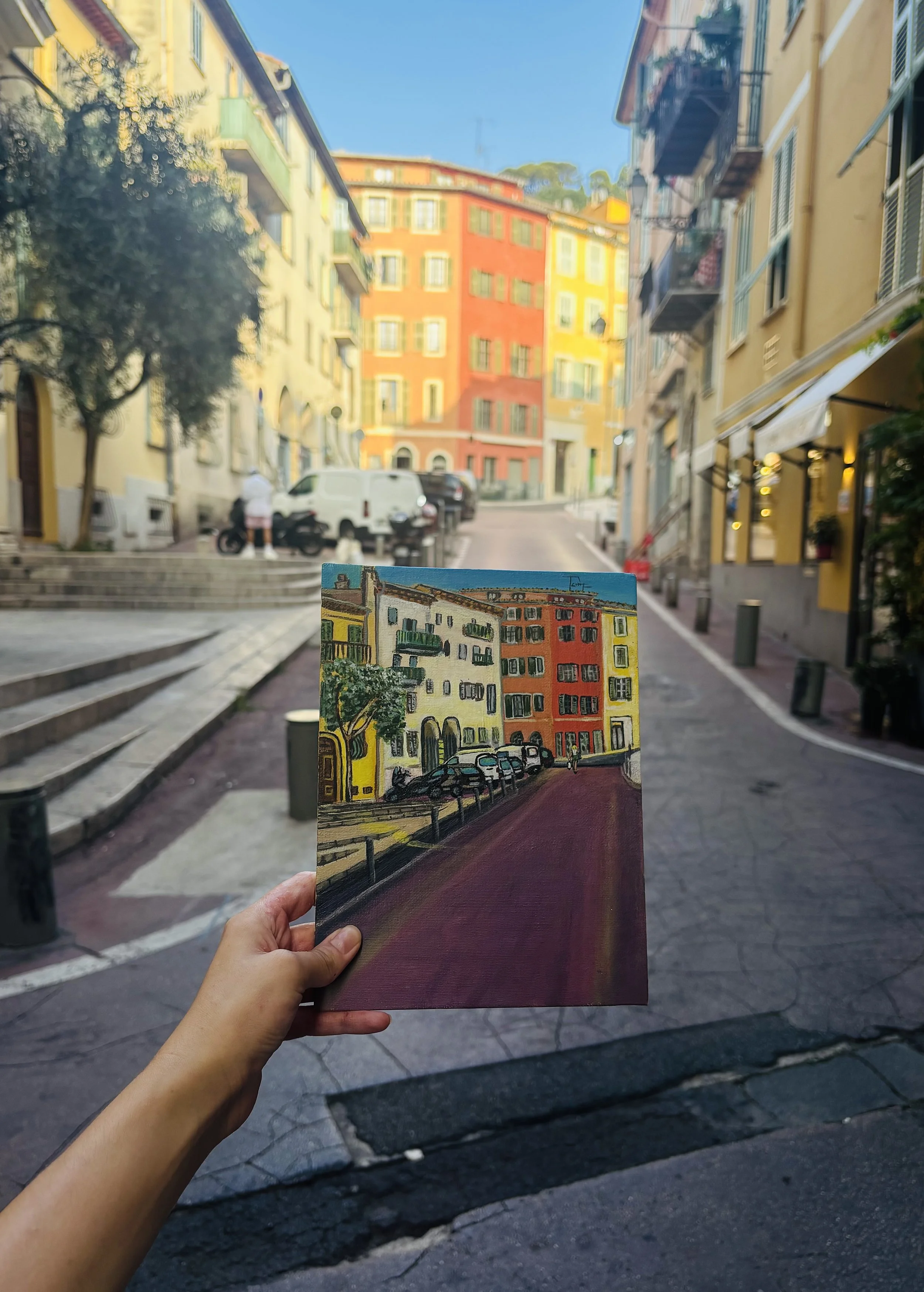 A person is holding a small painting of a colorful city street scene in front of the actual street, creating a visual comparison.