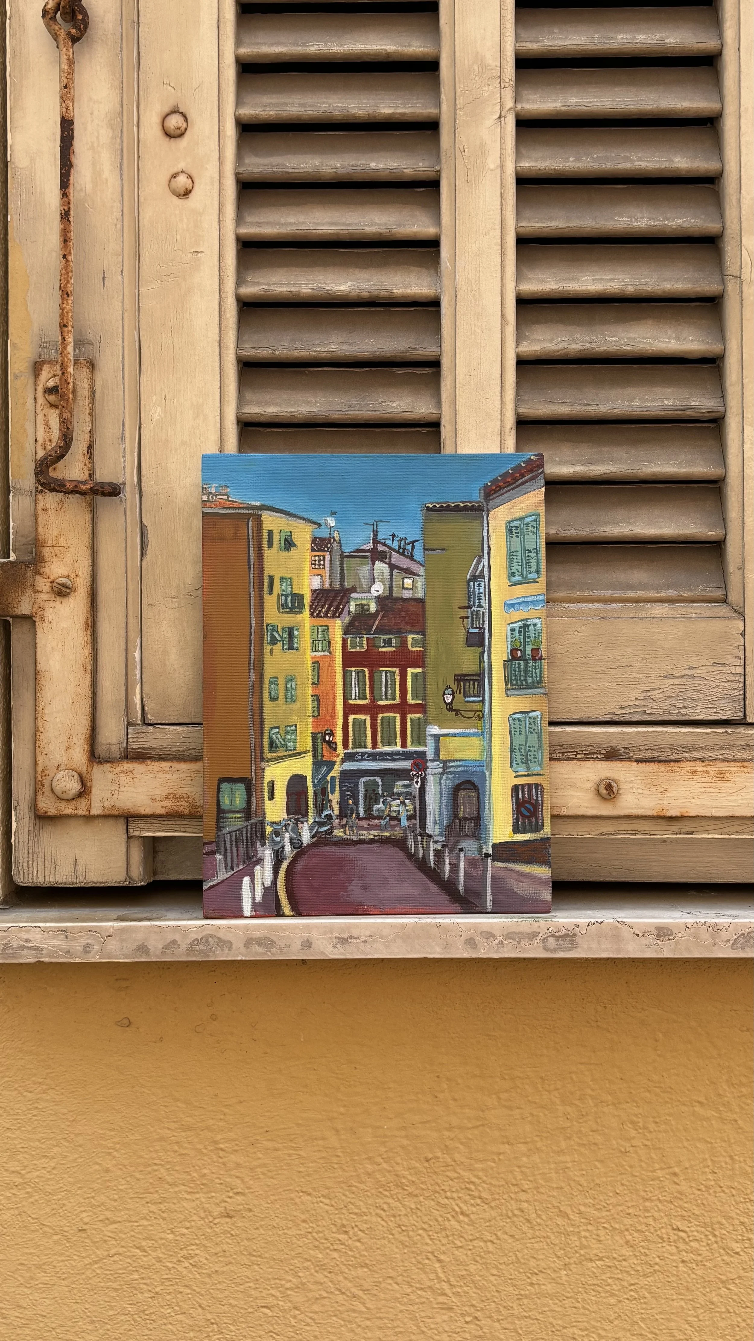 A colorful painting of a European city street scene with tall buildings, shops, and pedestrians.