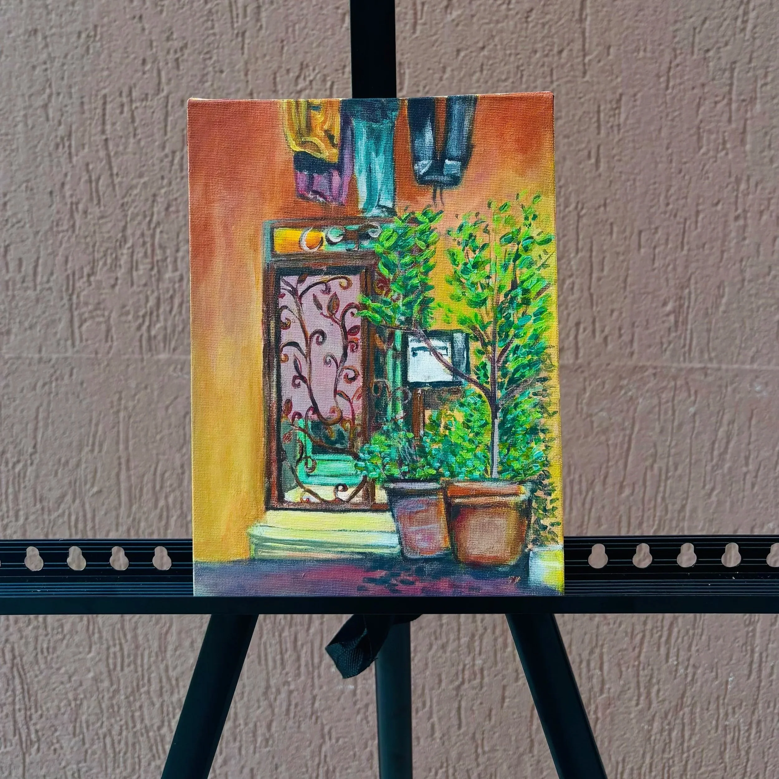 A colorful painting of a window with foliage outside, placed on a stand with black legs, against a beige textured wall.