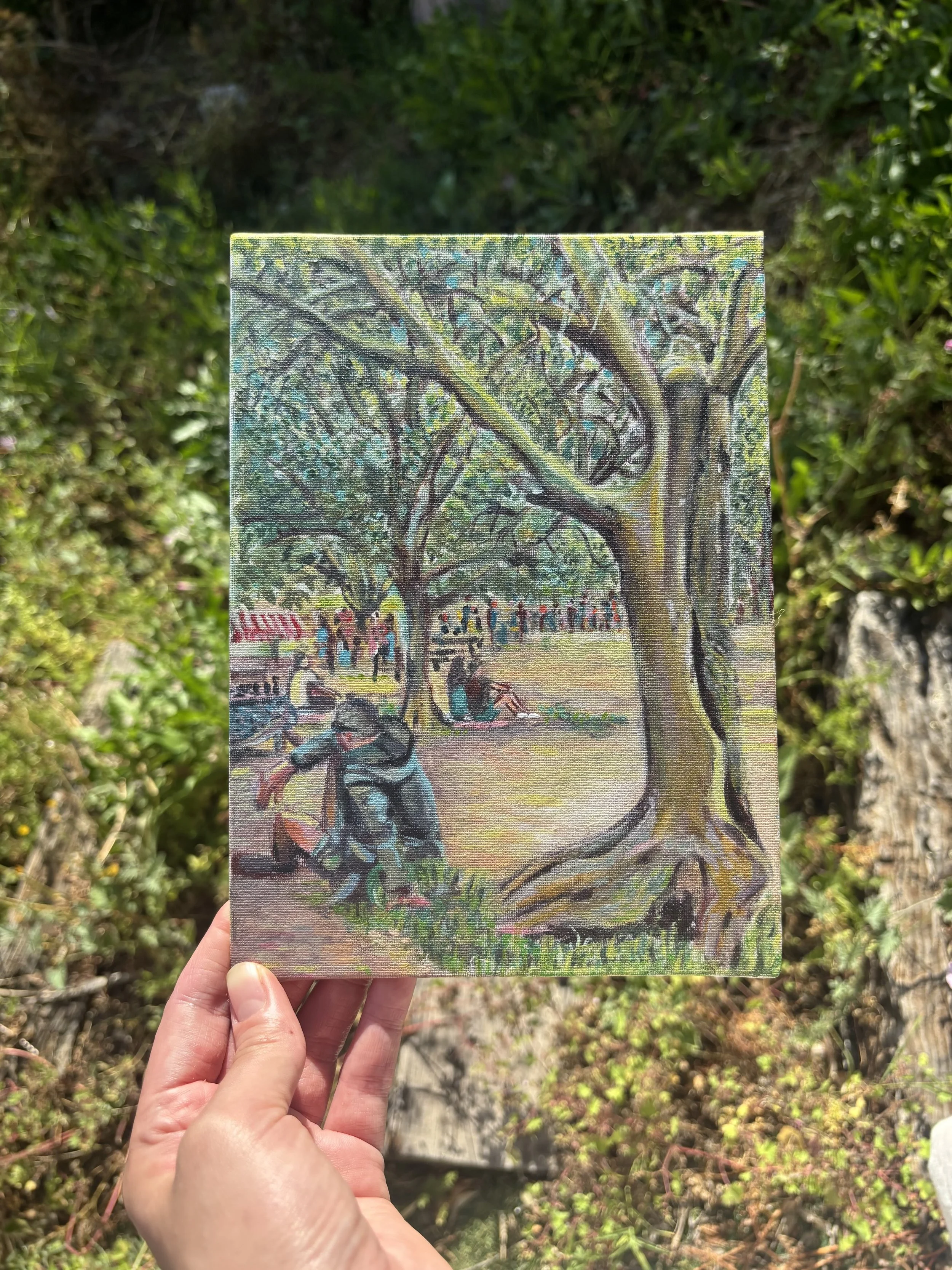 A hand holding a small painting of a tree with people underneath, set outdoors with green foliage in the background.
