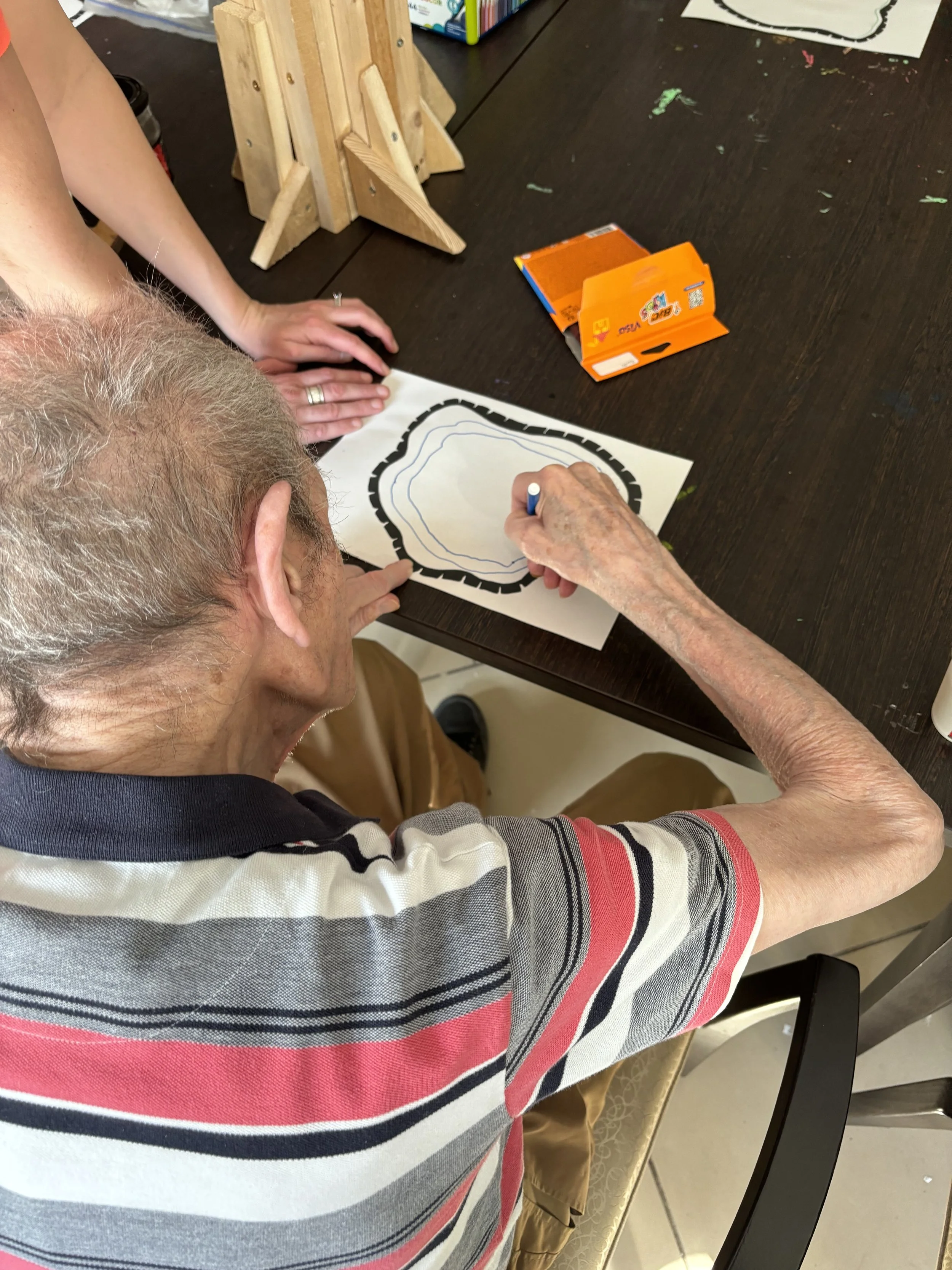 An elderly person sitting at a table, tracing a topographical map with a blue marker, while another person guides their hand. The table has a cardboard box, a wooden structure, and some art supplies. There are books on a shelf in the background.