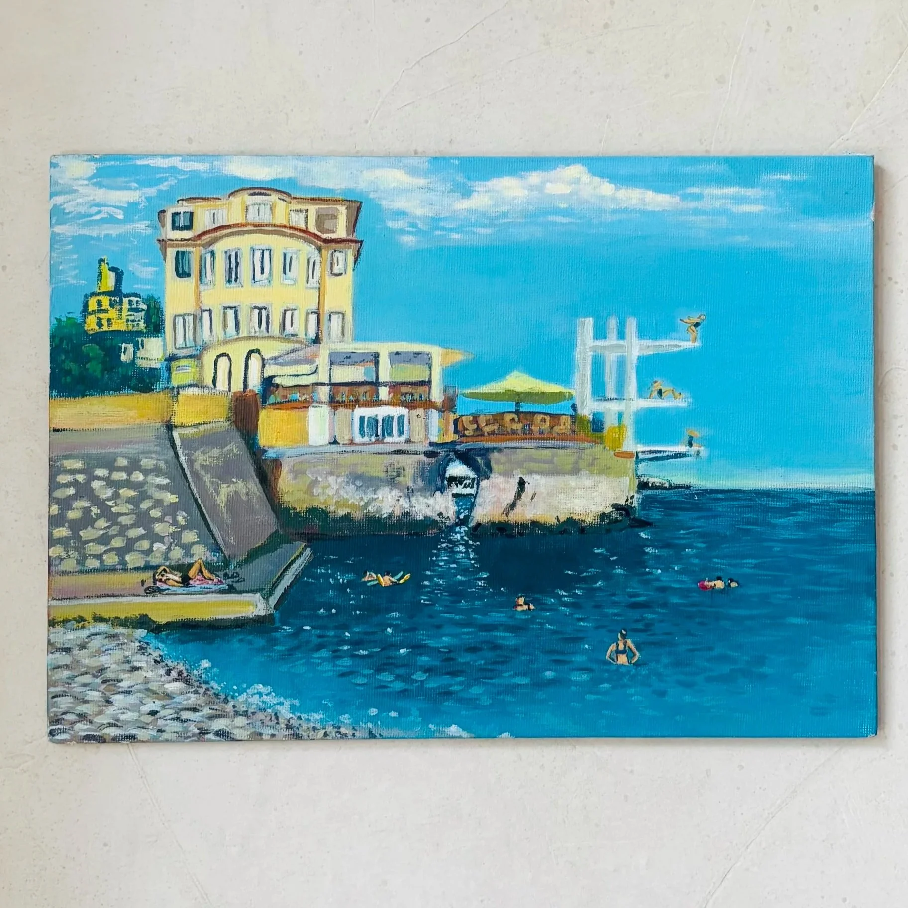 A colorful painting of a seaside scene with a yellow building, people sunbathing and swimming, and a blue sky with white clouds.