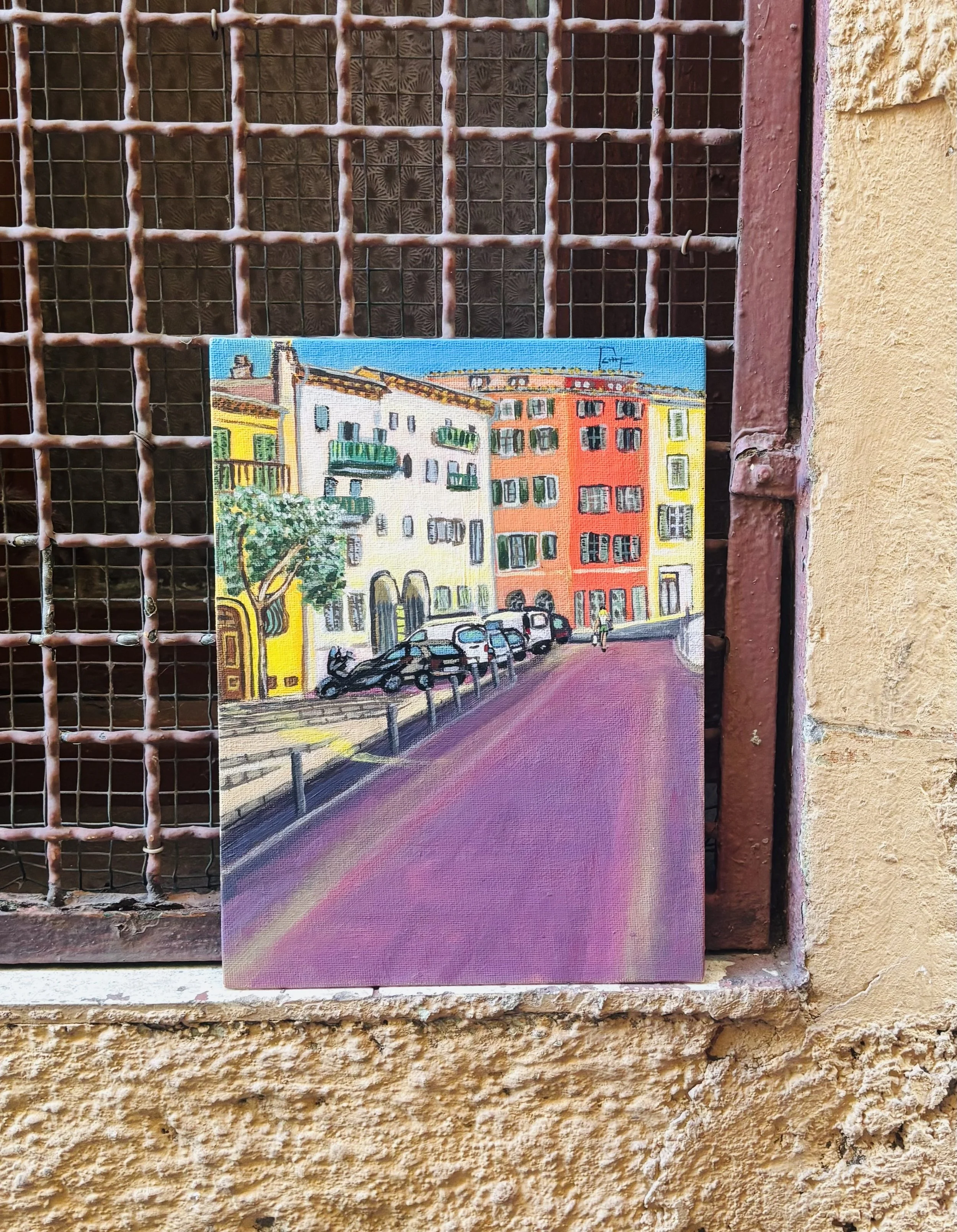 Colorful painting of buildings with parked cars, trees, and people on a city street, displayed on a textured yellowish wall and a rusty metal grid.