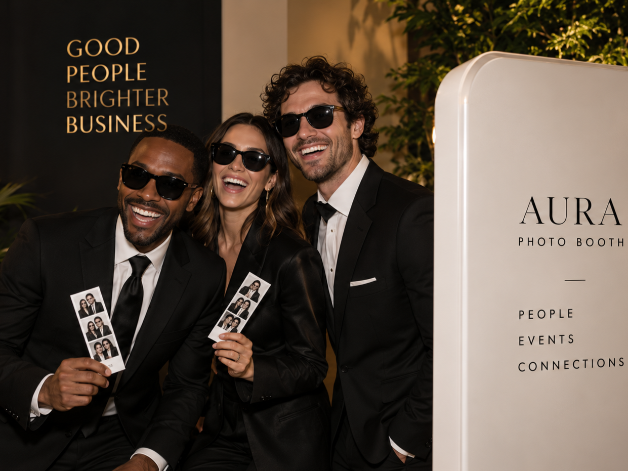 Three people in black suits and sunglasses smiling and holding photo booth strips at Aura photo booth event, with a sign in the background that reads 'Good People Brighter Business.'