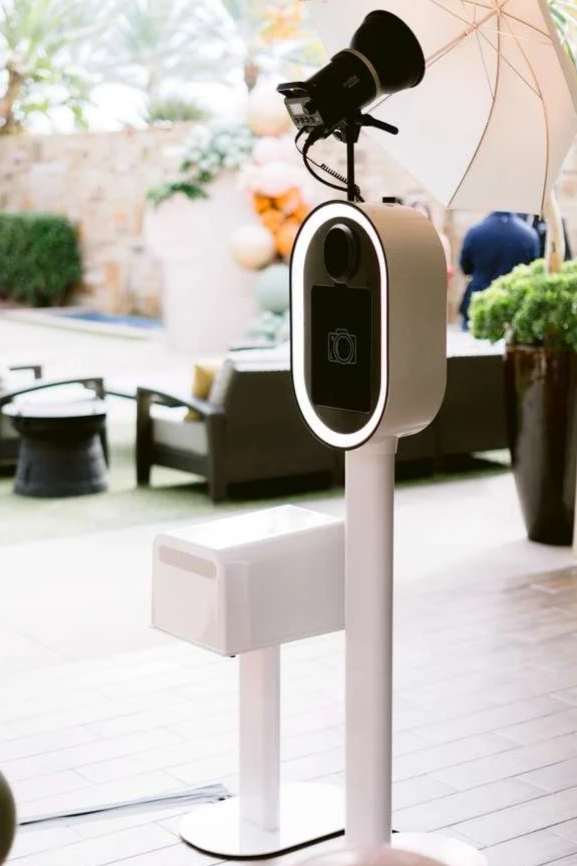 A contactless temperature scanning kiosk with a built-in camera and a spotlight, set up outdoors with seating and decorative plants in the background.