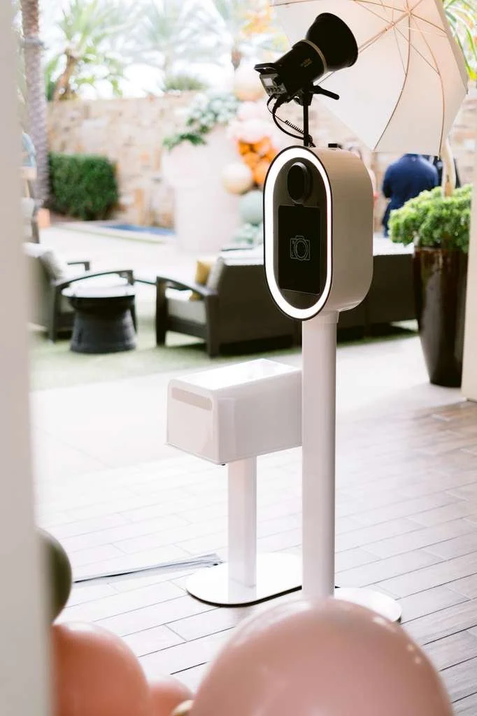 Photo of a photo booth station set up outdoors with a white umbrella, a camera mounted on top, and a screen for taking pictures. In the background, there are outdoor furniture and potted plants.