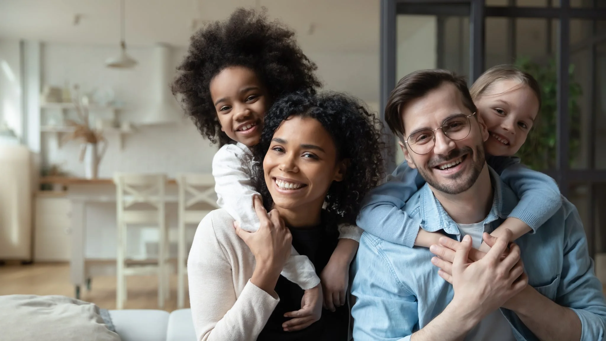 Family of four smiling and playing together in their home. The mother and father are holding two children, one on each parent's shoulders, in a cozy living room with natural light.