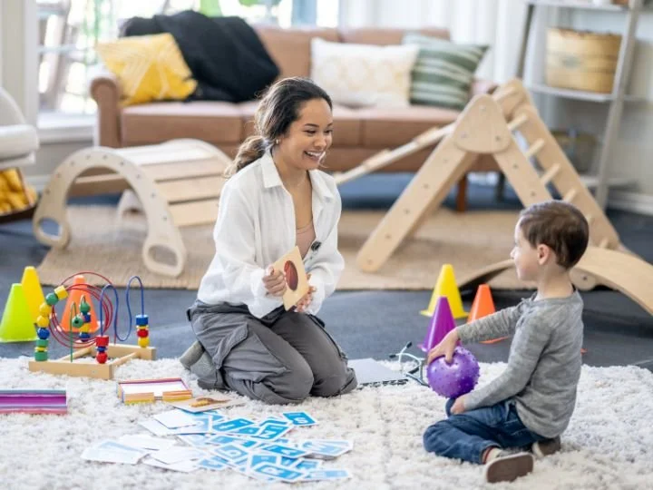 A woman and a young boy playing with toys and cards on a carpet in a bright living room with a wooden climbing structure.