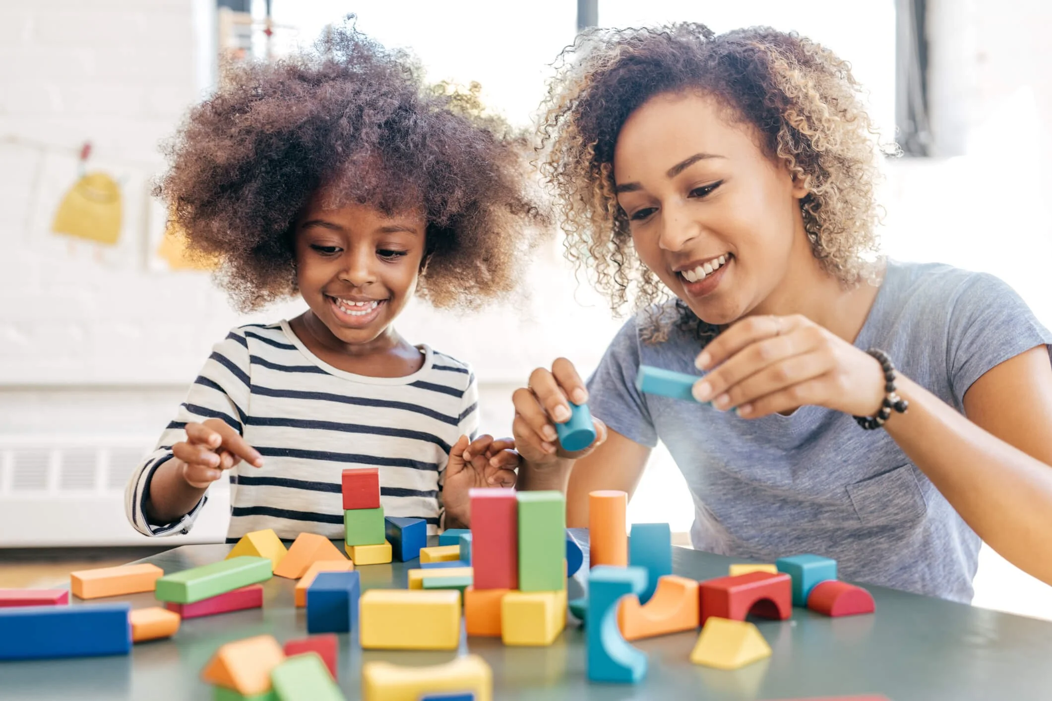 A woman and a girl playing with colorful wooden blocks on a table in a bright room.