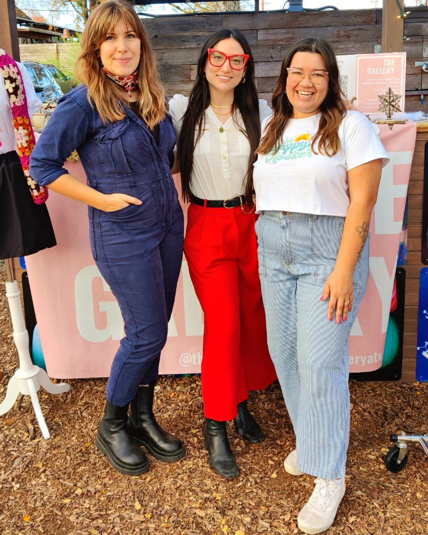 Three women standing outdoors at an event, smiling, with a booth behind them displaying signs and artwork.