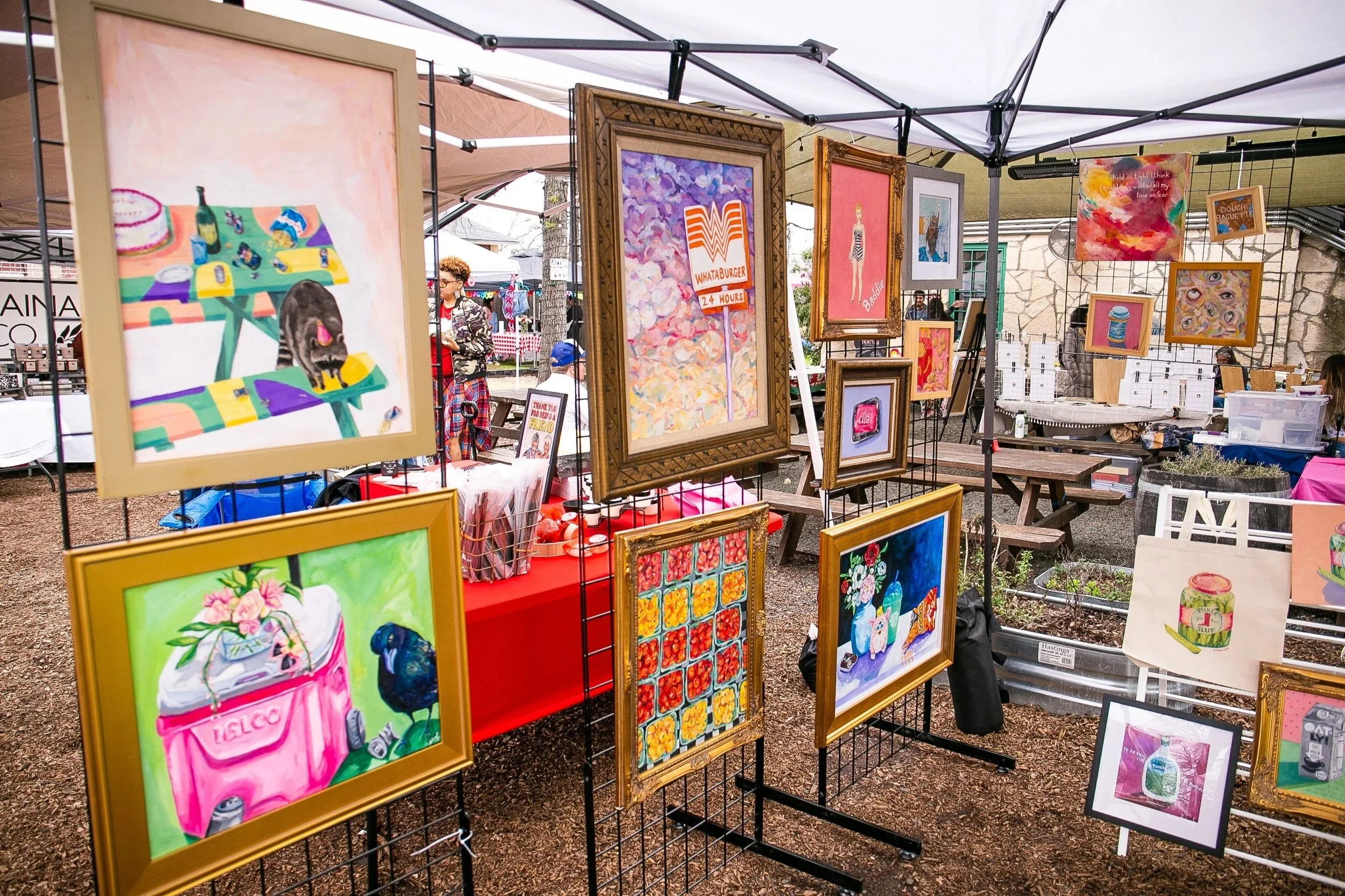 An outdoor art market displaying colorful paintings on a metal grid and frames, with some artwork featuring flowers, abstract designs, and pop culture references. People are browsing and engaging with the art under tents.