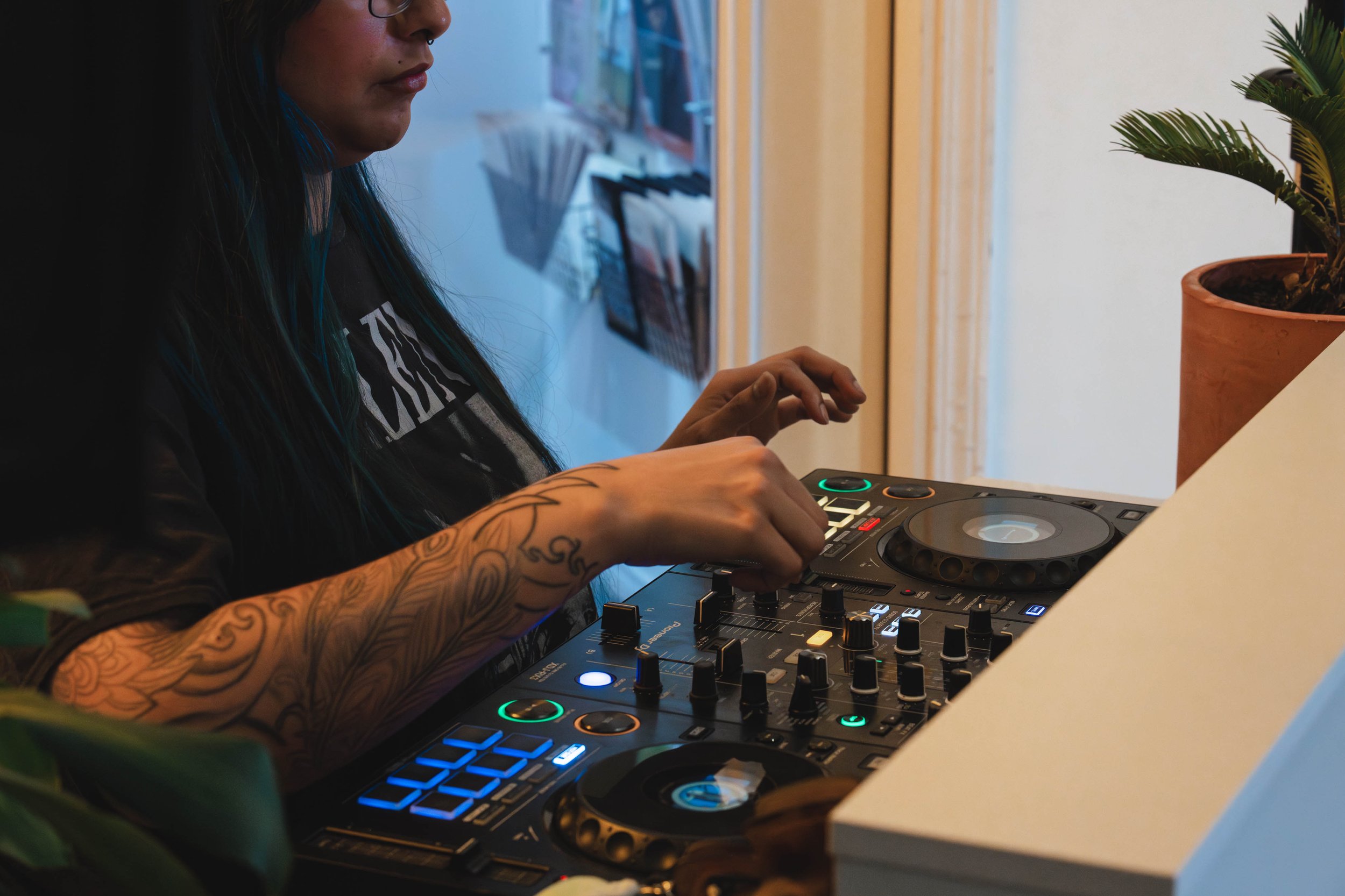 A person with long dark hair and tattoos on their arm is using DJ equipment, including a mixer and turntables, near a potted plant on a white surface inside a room.